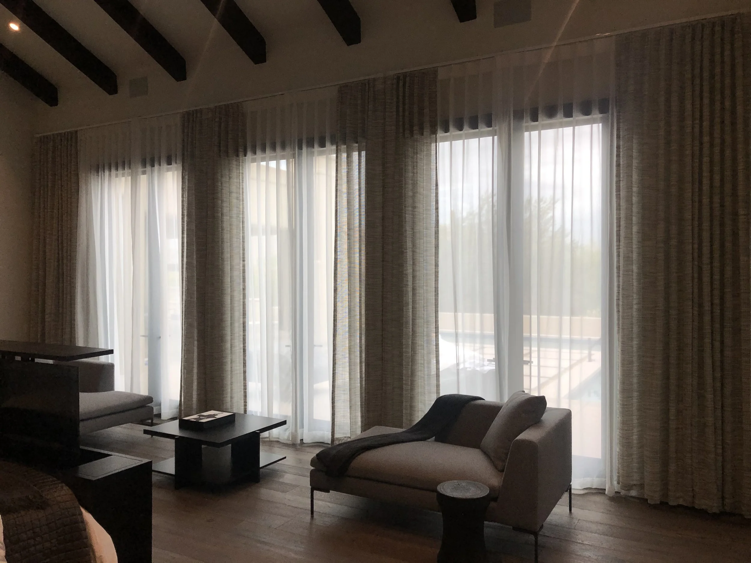 Living room with large windows covered by sheer white curtains and heavier patterned drapes, with lounge chairs and coffee table.