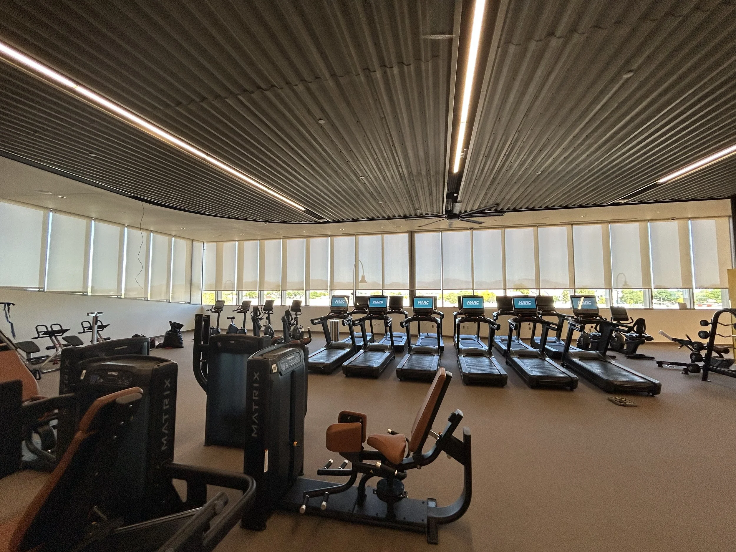 Indoor gym with treadmills facing large windows, ceiling with wooden slats, and exercise bikes in the foreground.