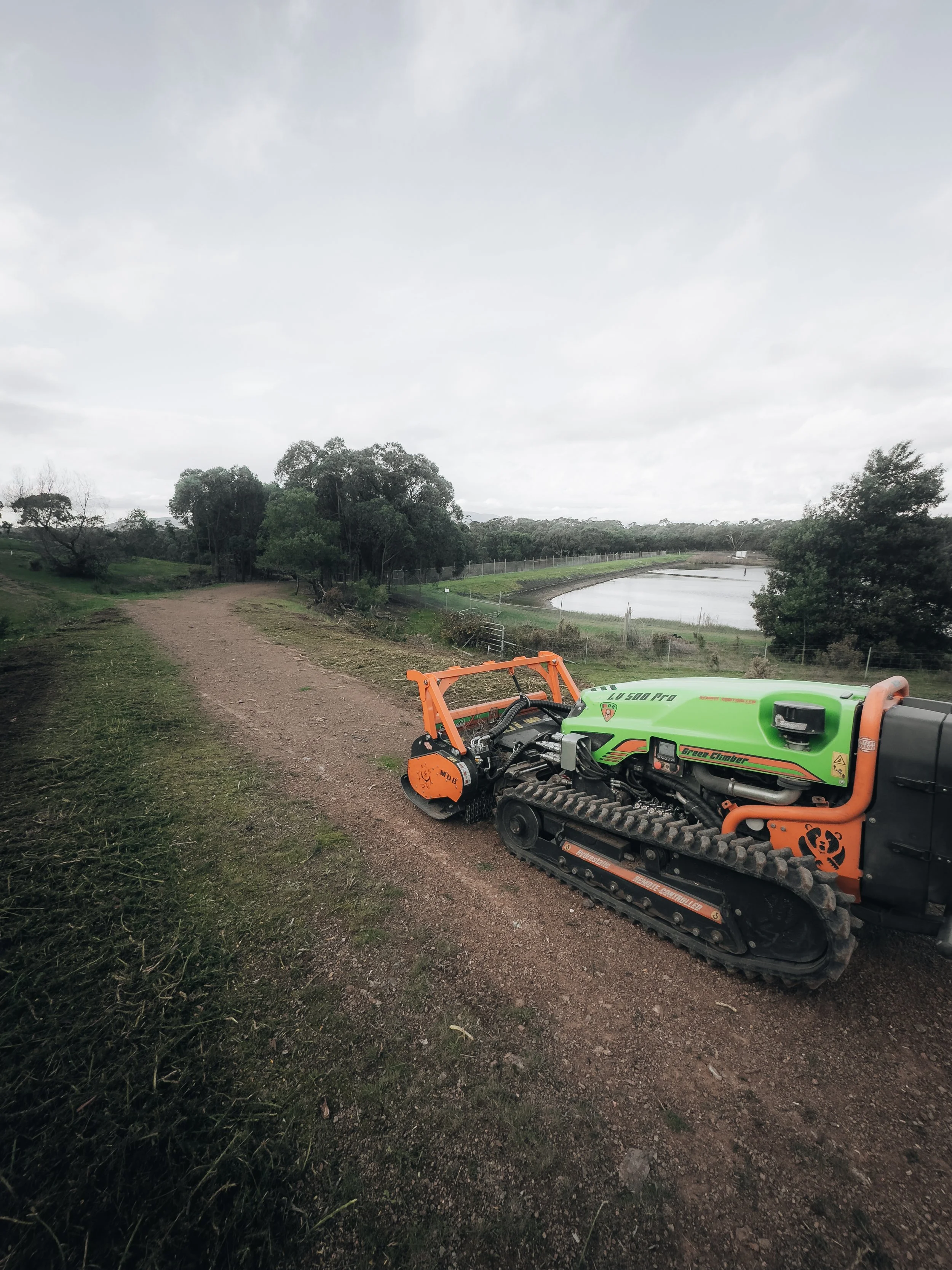 A green and orange tracked outdoor machinery on a dirt path near a body of water surrounded by trees.