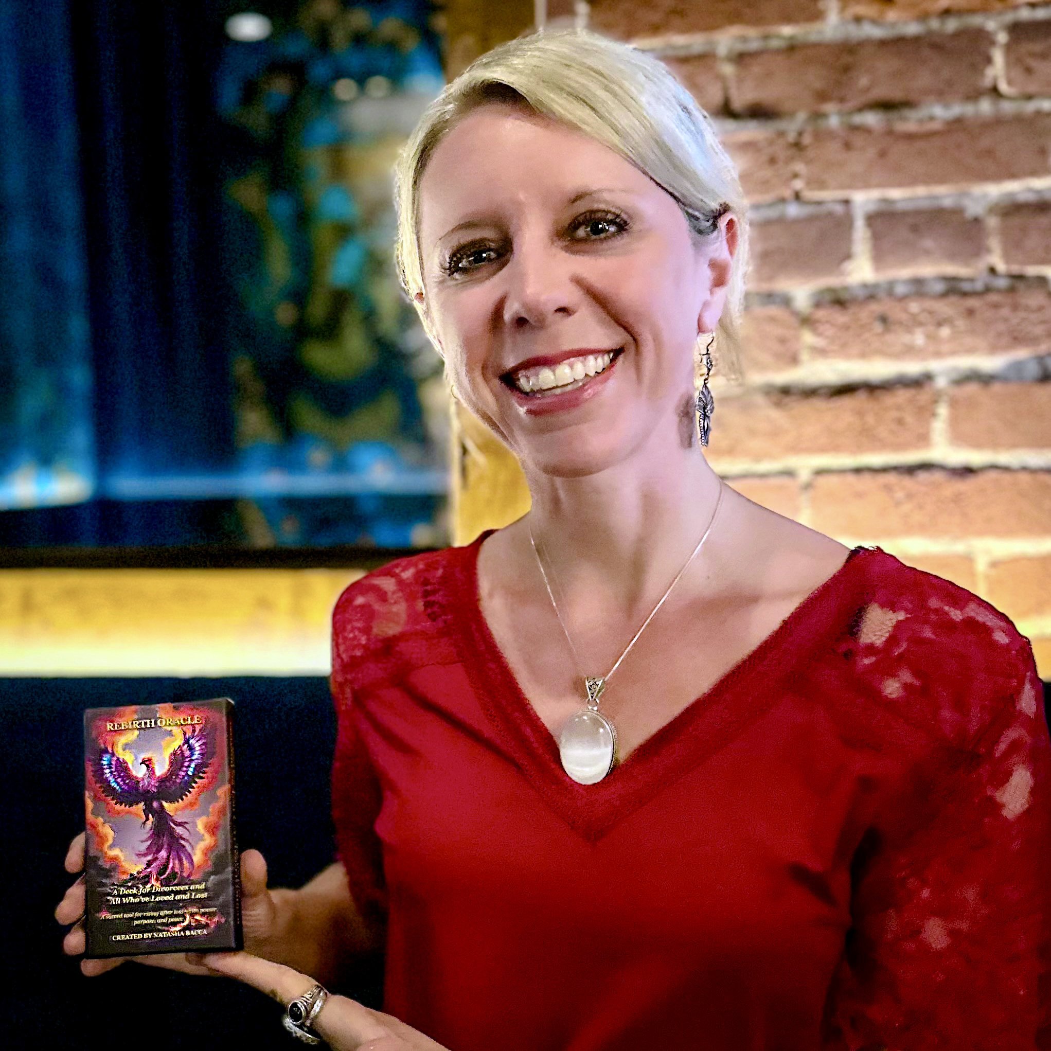 A woman with blonde hair smiling and holding a tarot card titled "Rebirth Oracle" in her right hand. She is wearing a red dress and jewelry, including earrings, a necklace, and a ring. The background shows a brick wall and some colorful decor.