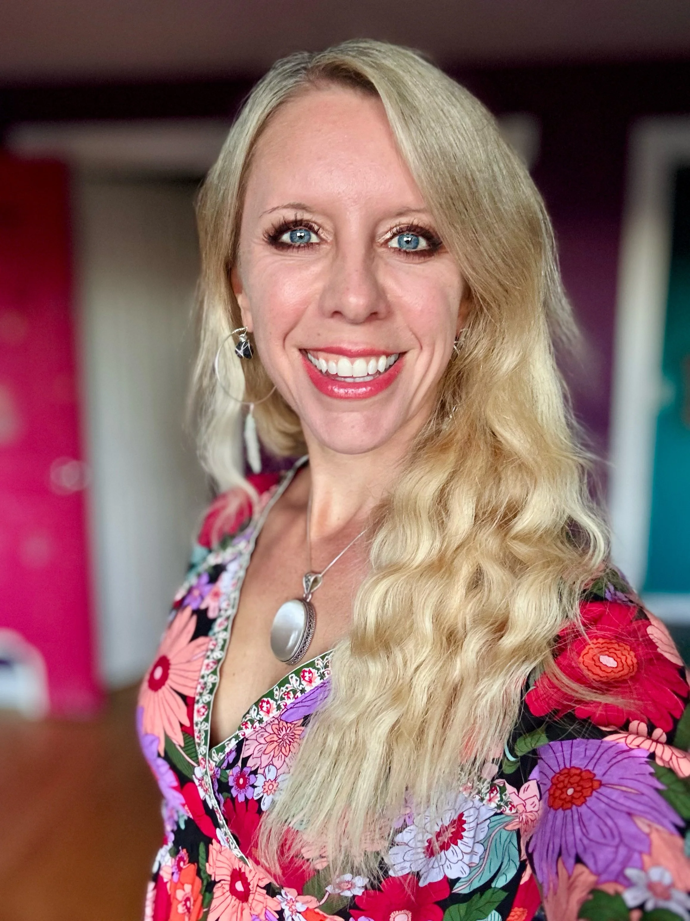 Close-up of a smiling woman with long blonde hair, blue eyes, and wearing a colorful floral dress.