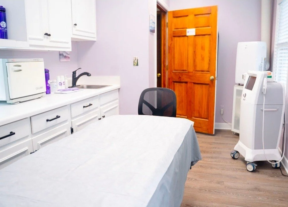 A room with a table covered by a white tablecloth, a black office chair, a closed wooden door, white cabinets, a microwave, and medical equipment.