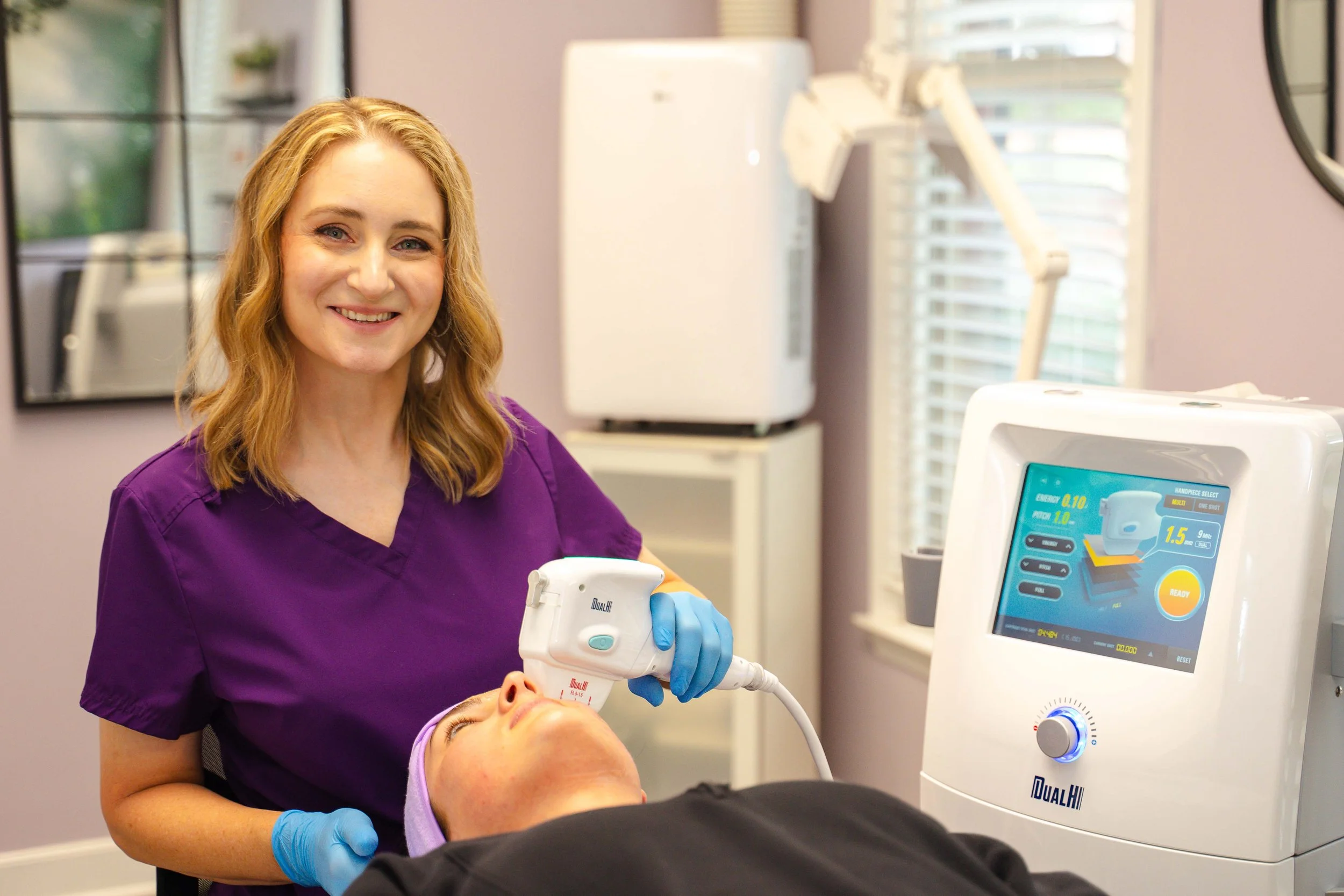 A female healthcare professional with blonde hair, wearing purple scrubs and blue gloves, is performing a medical procedure on a patient lying down. The patient is connected to a medical device with a touchscreen display showing various metrics, in a clinical setting with medical equipment and windows with blinds in the background.