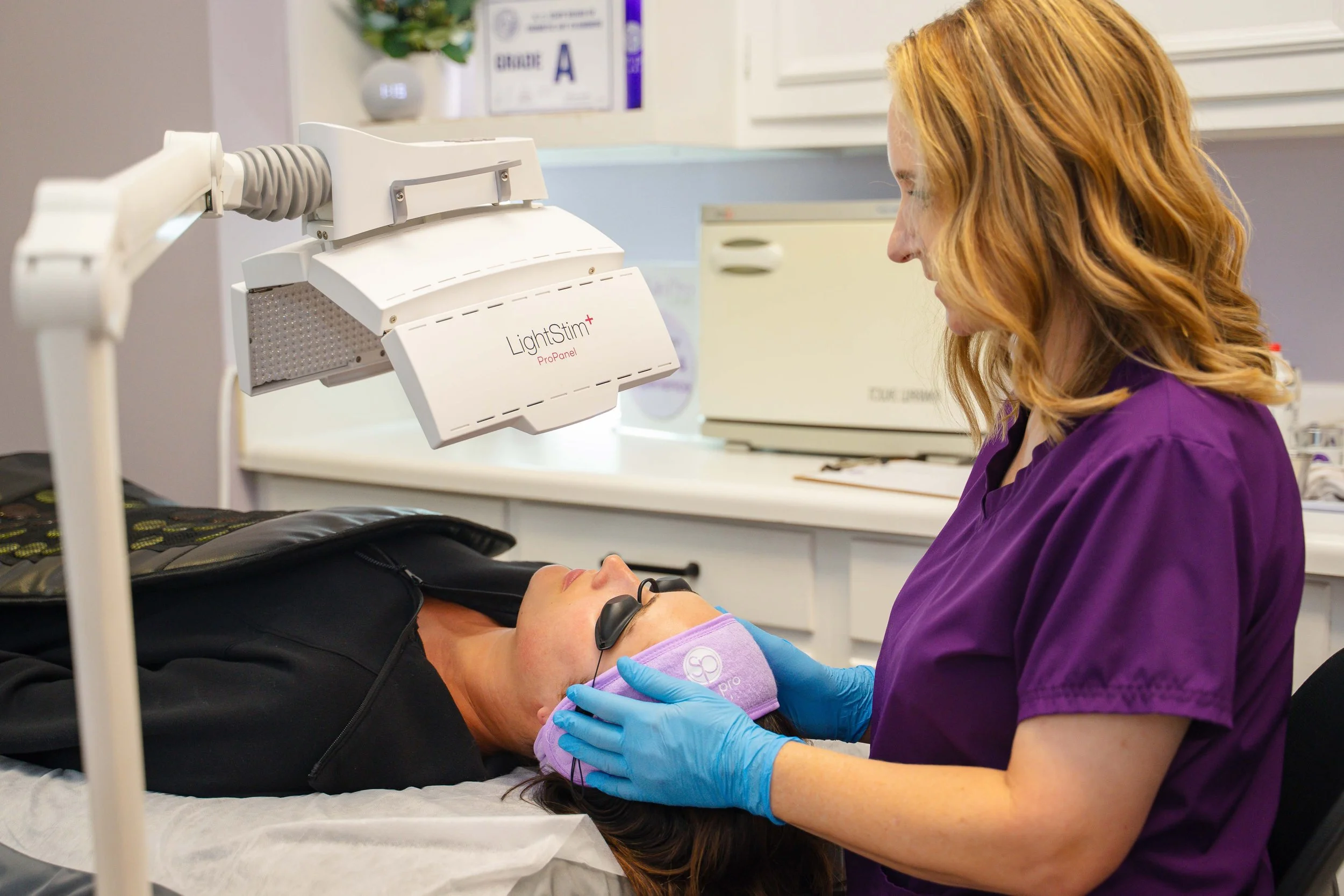 A woman with glasses and a purple medical uniform is administering a procedure to a patient lying on a medical bed. The patient is wearing protective goggles and has her head wrapped in a purple cloth. Medical equipment, including a LightStim device, is visible in the background.