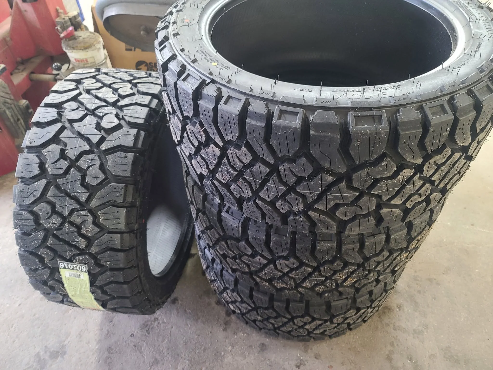 Four large off-road tires stacked on a workshop floor with red tool chest and workbench in the background.