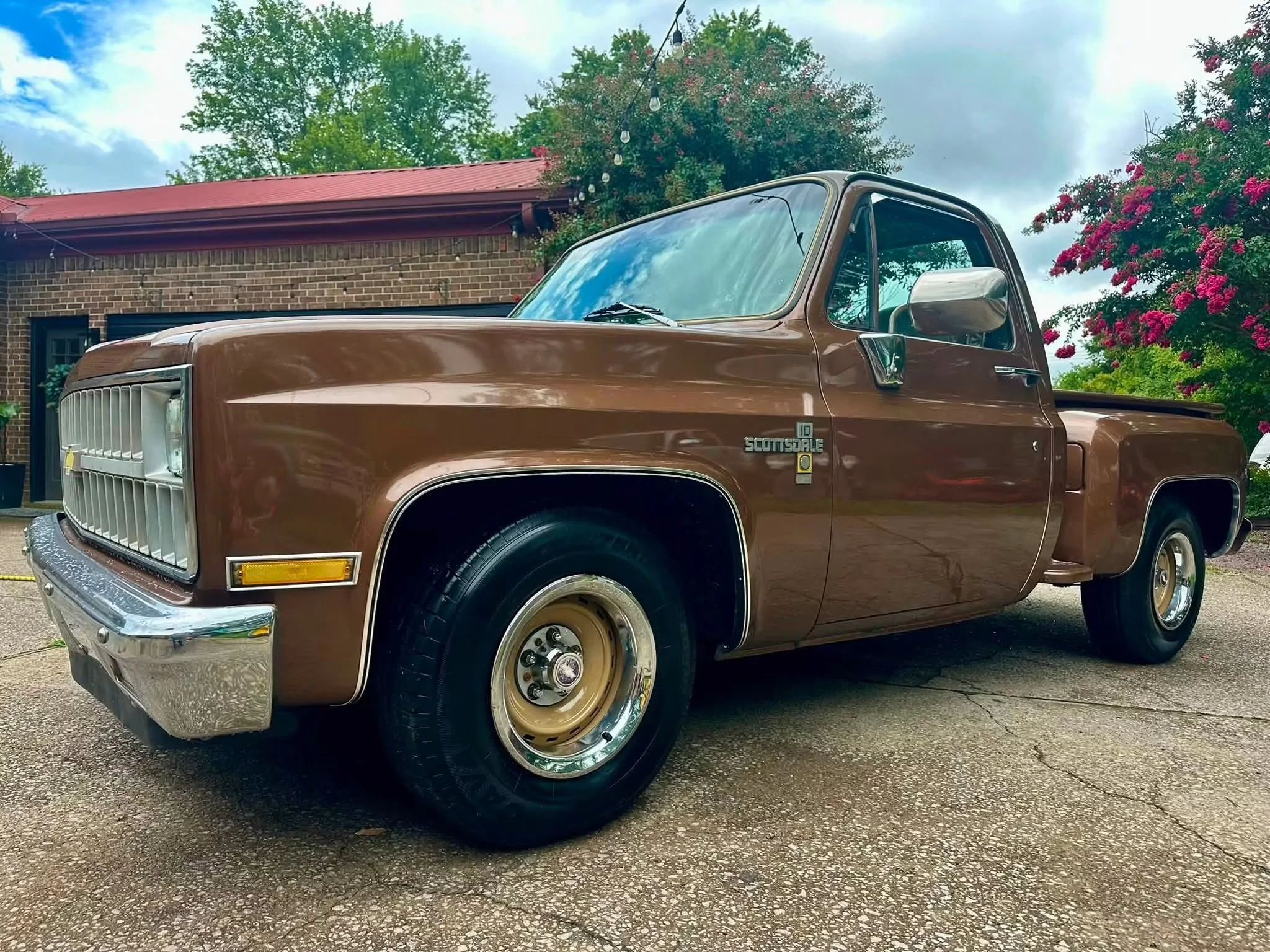 A vintage brown pickup truck parked outdoors in front of a brick building with a red roof. The truck has a chrome bumper and beige wheels. There are trees and flowering bushes in the background, and string lights hanging overhead, suggesting a festive or decorative setting.