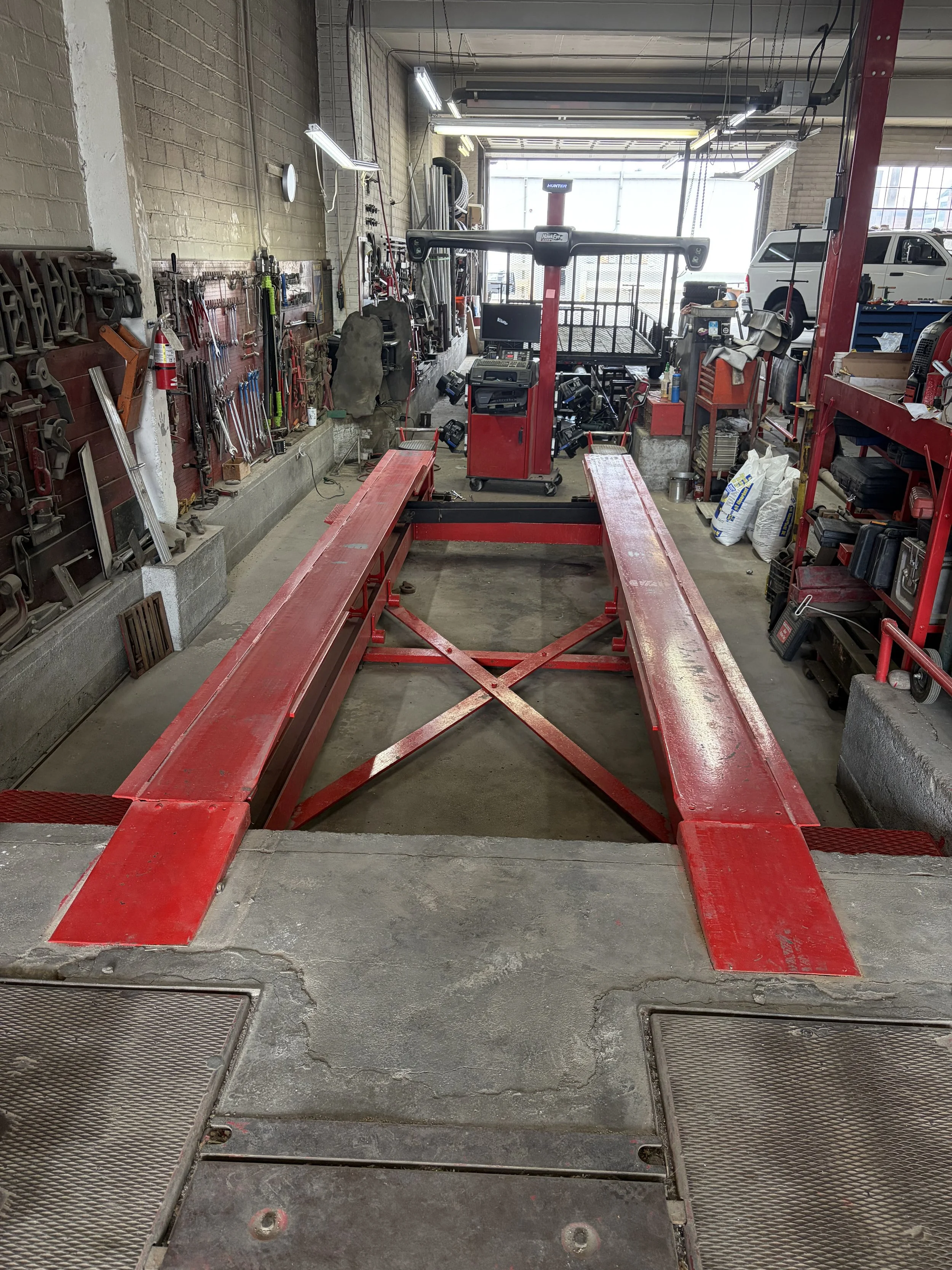 Inside an automotive workshop with a red vehicle lift frame in the center, tools hanging on the wall to the left, and various equipment and supplies scattered around, including a white vehicle parked in the background near the garage door.