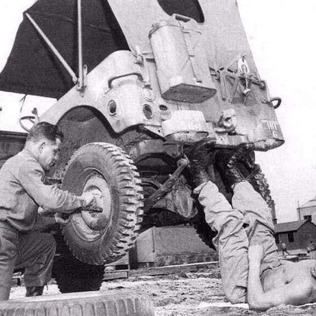 Two men working under a large military truck; one man is checking or repairing the truck's wheel, the other is lying underneath the vehicle, possibly inspecting or repairing it, with a parasol in the background.
