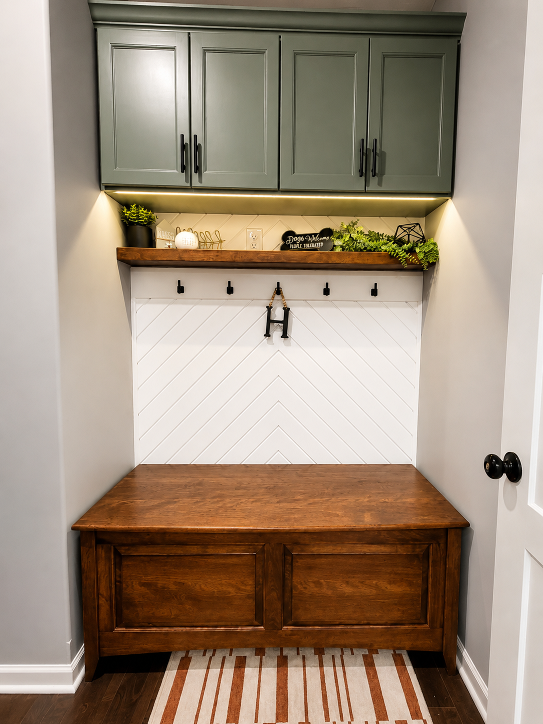 A small entryway with a wooden bench, black hooks on a white paneled wall, and green cabinets with decorative plants and signs on a wooden shelf.
