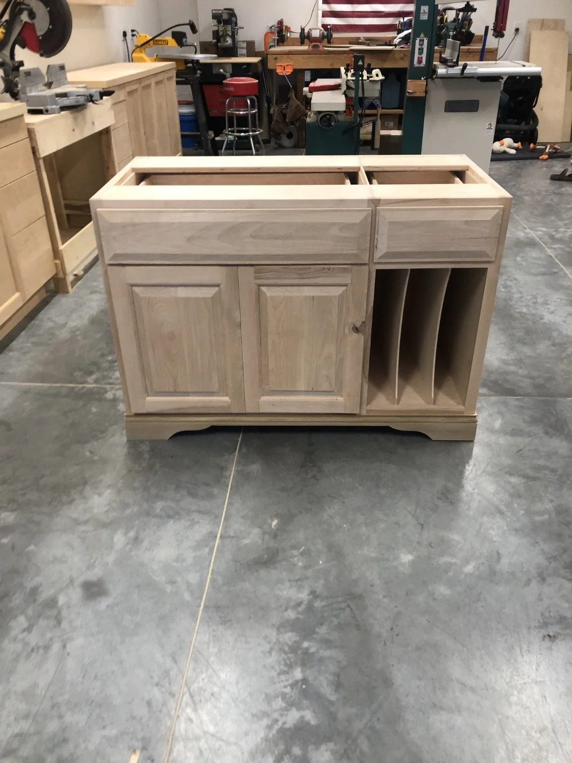 Unfinished wooden kitchen cabinet with compartments for a sink and wine bottles, in a workshop.