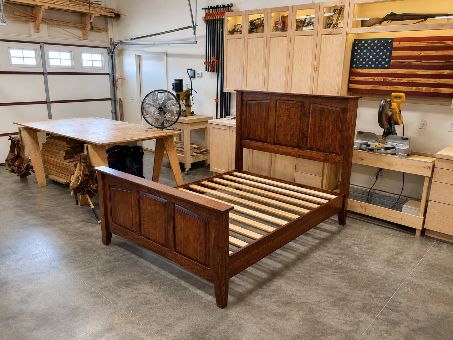 A woodworking workshop featuring a partially assembled wooden bed frame, a large work table, various woodworking tools, clamps, and storage cabinets, with an American flag wall decoration.