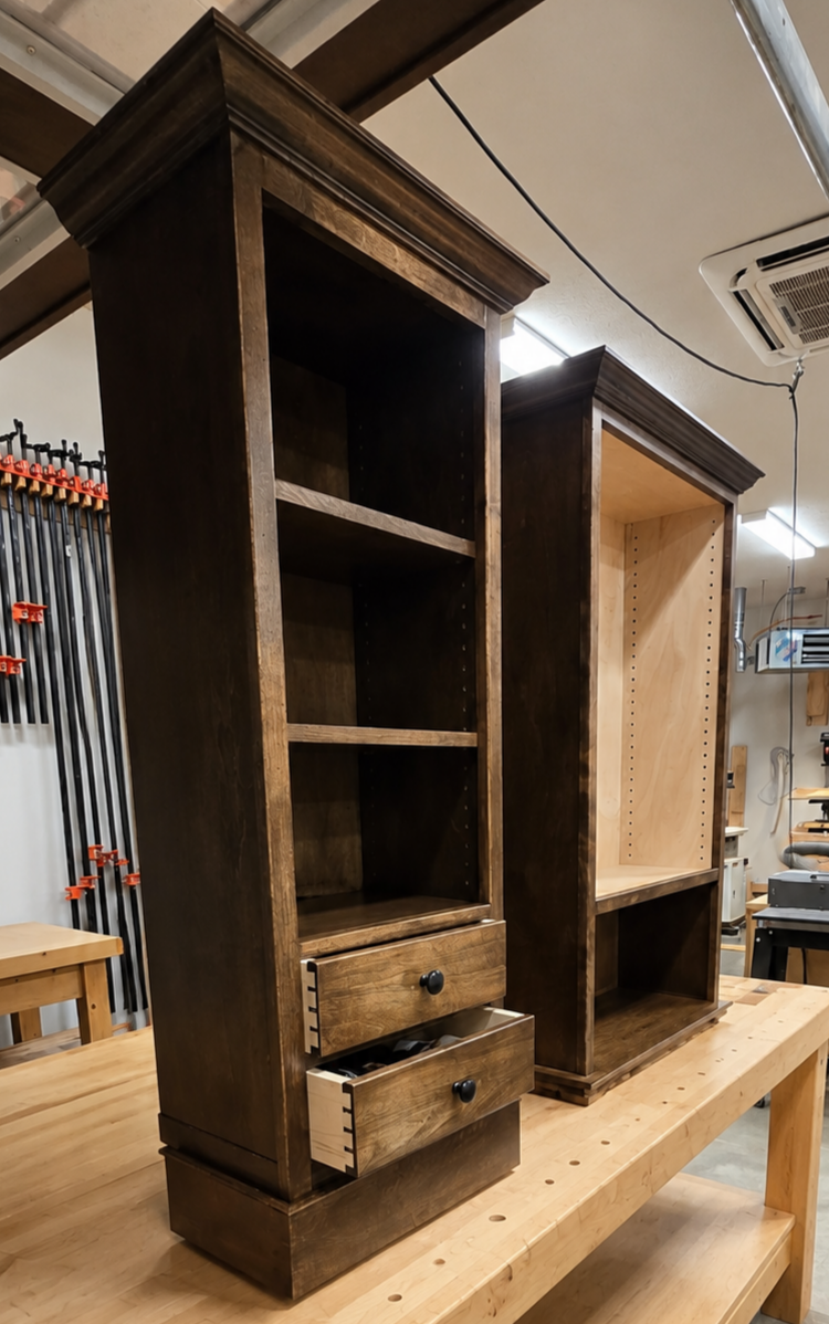 Two wooden bookshelves in a workshop, one dark stained with drawers at the bottom, the other unfinished with adjustable shelving.