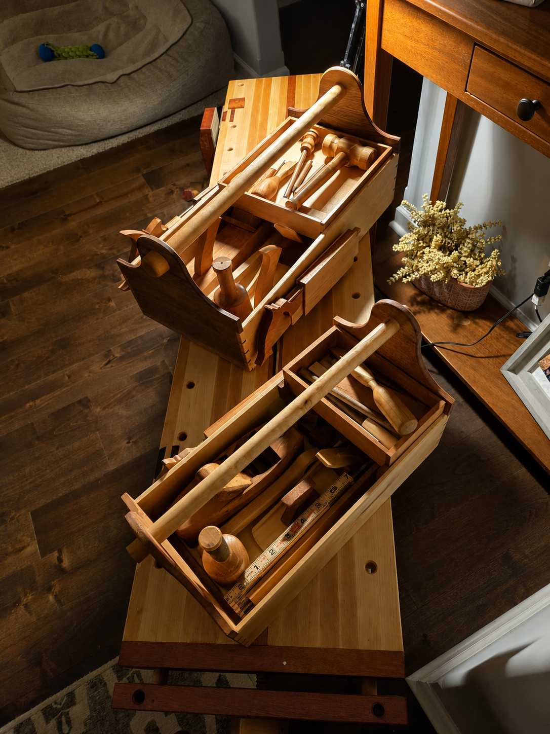 Multiple wooden pestle and mortar sets and measuring tools stored in wooden boxes on a bench in a room with hardwood floor. A dog bed and a potted plant are also visible.