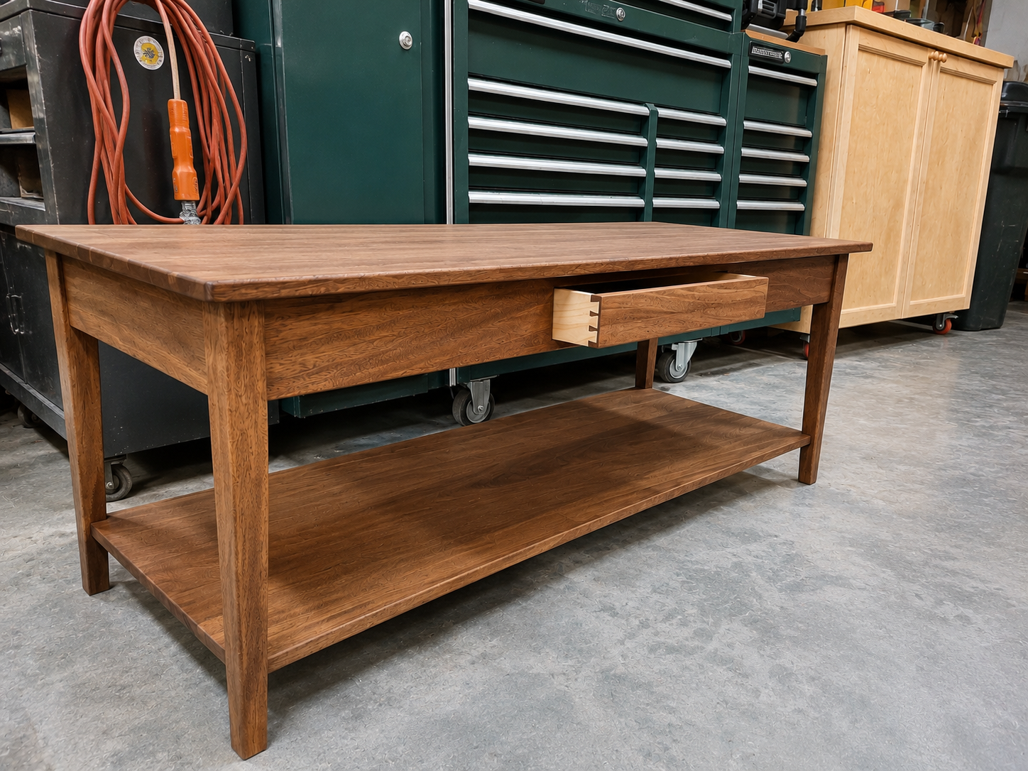 A wooden workbench with a drawer and lower shelf, in a workshop setting with tools and storage cabinets in the background.