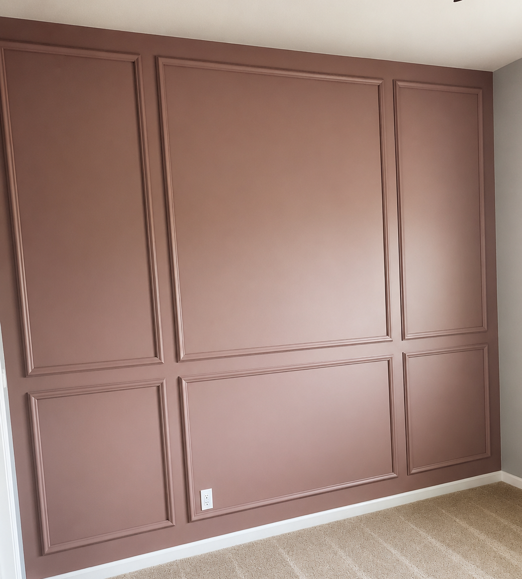 A wall with decorative wainscoting panels painted in a muted pink color, with beige carpet flooring and a white electrical outlet near the bottom.