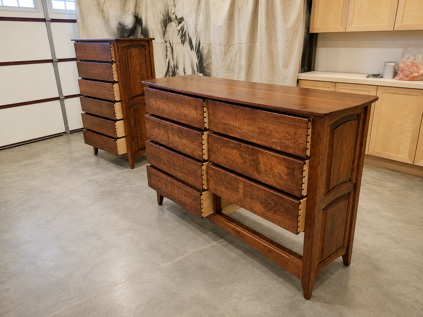Two wooden dressers with six drawers each in a garage or workshop space, with a beige curtain, a countertop with a plastic bag and other items in the background.
