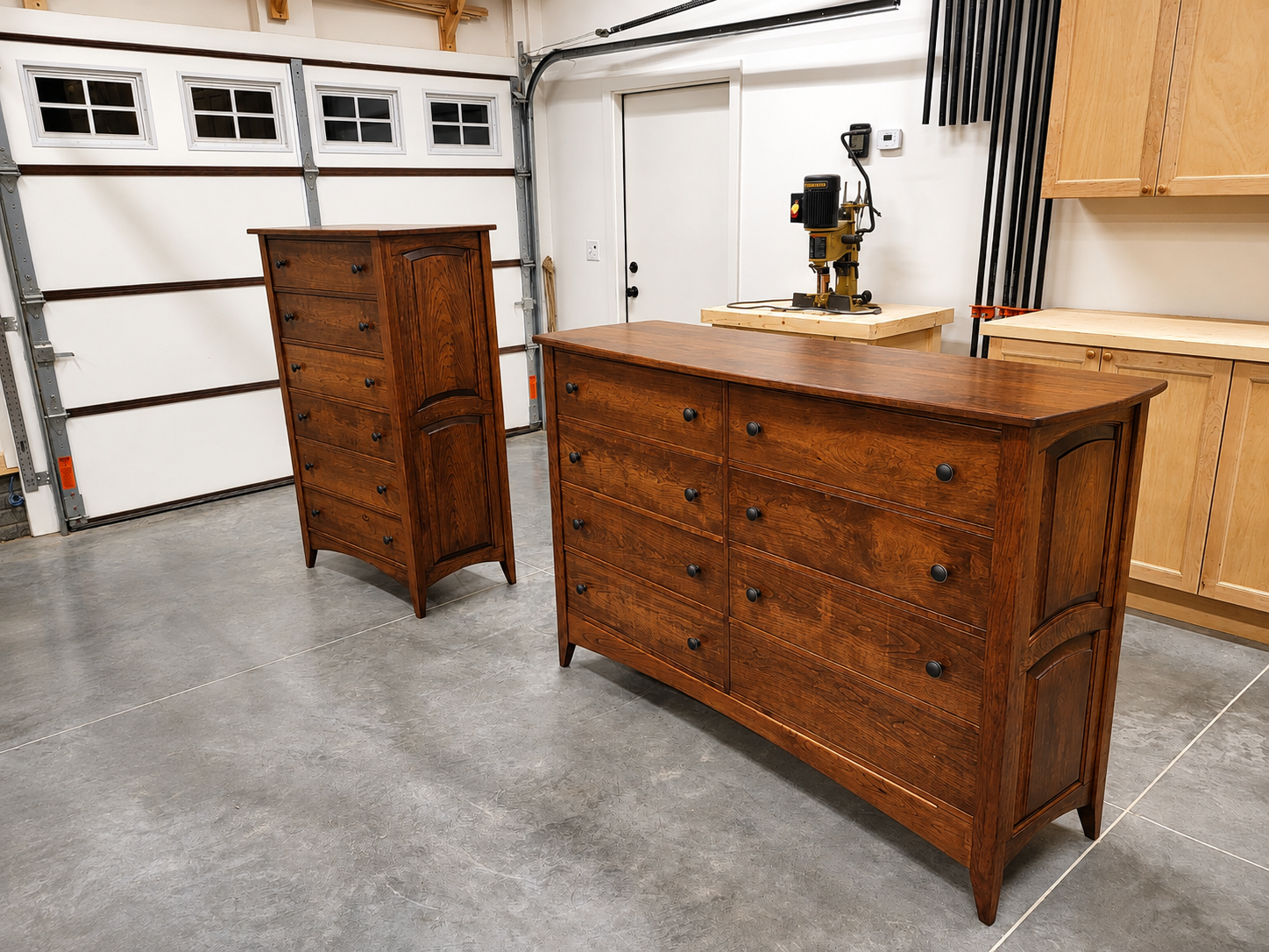 A garage with two wooden dressers, a tall chest of drawers and a wide dresser with multiple drawers, on a concrete floor, with a white garage door and a workbench with a power tool in the background.
