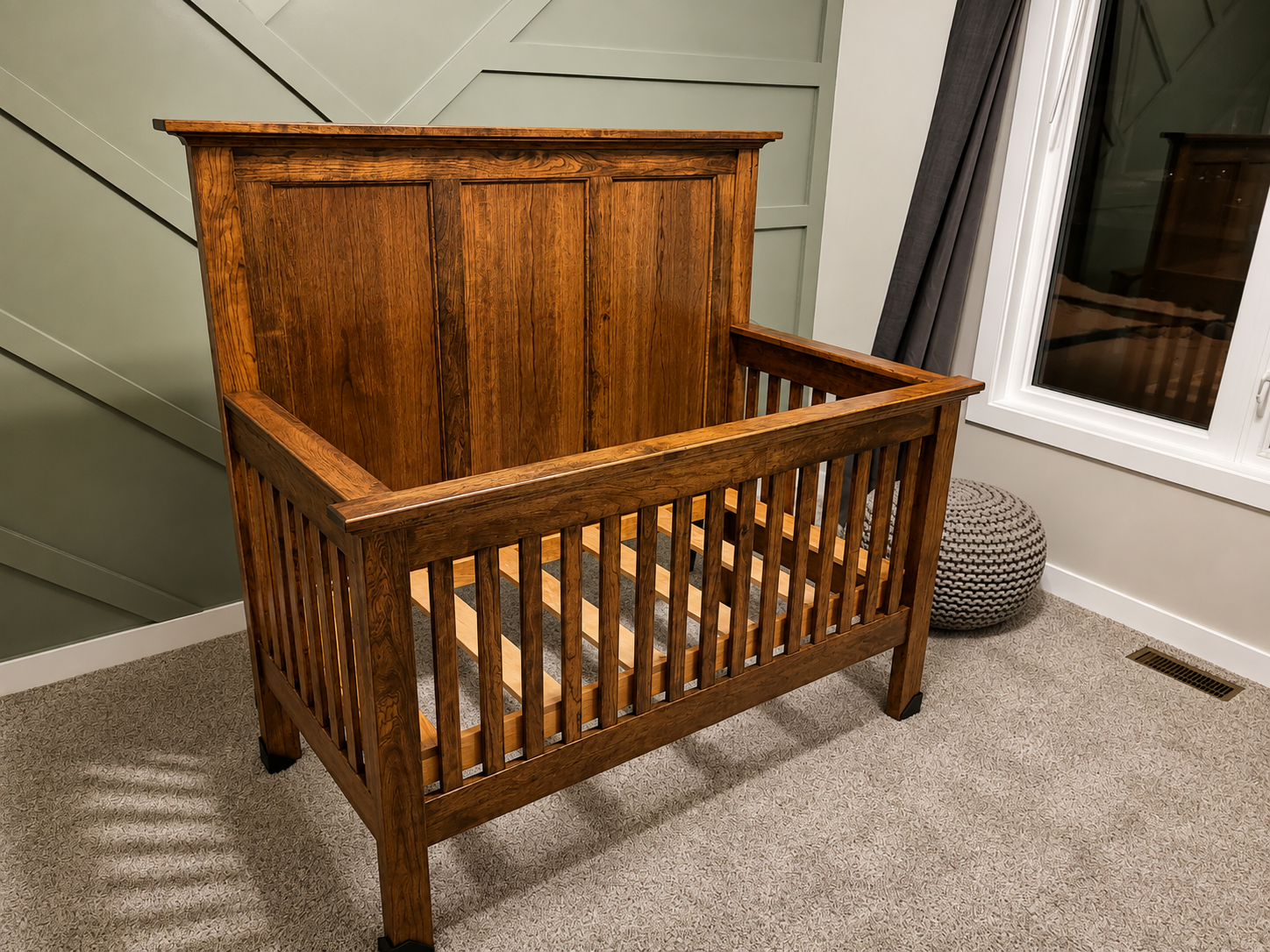 A wooden crib with slatted sides and a tall back panel in a room with beige carpet, a small round knitted pouf, a window with a dark curtain, and light-colored walls with decorative paneling.