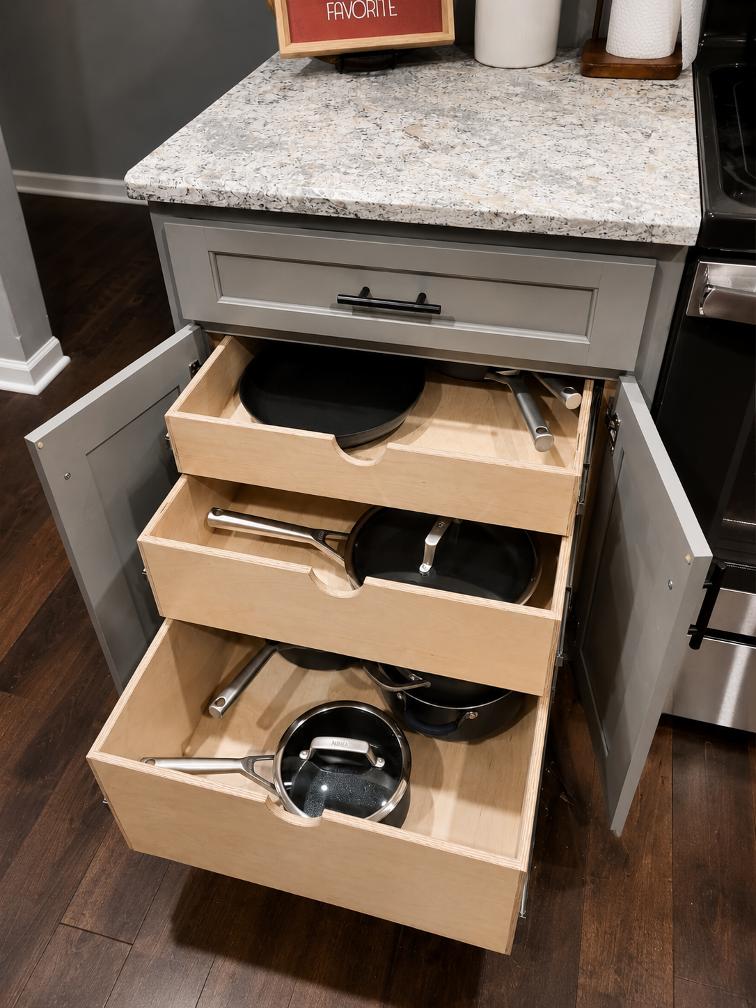 Open kitchen cabinet with three drawers containing pots and pans under a granite countertop.