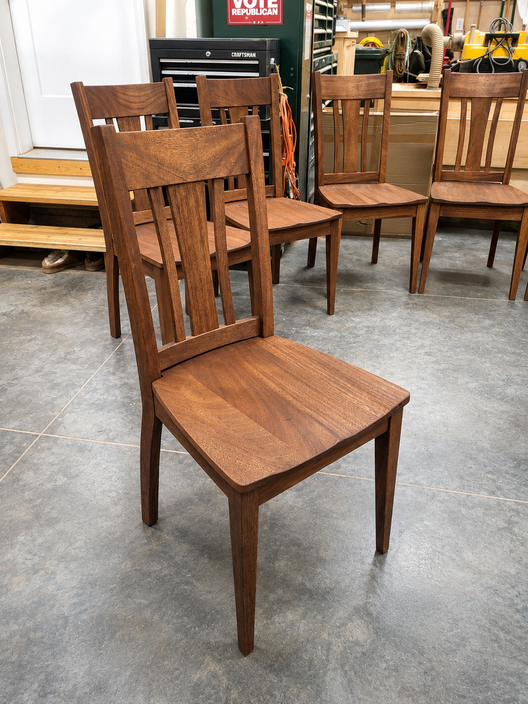 Set of four wooden chairs with slatted backs in a workshop, with various tools and equipment in the background.