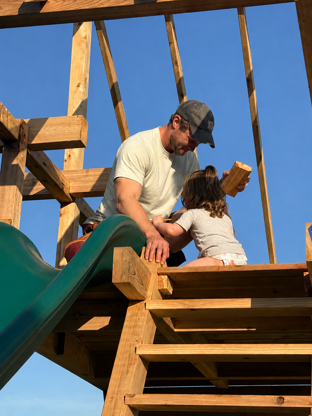 A man and a young girl on a wooden playground structure with a slide, building or fixing something together under a clear blue sky.