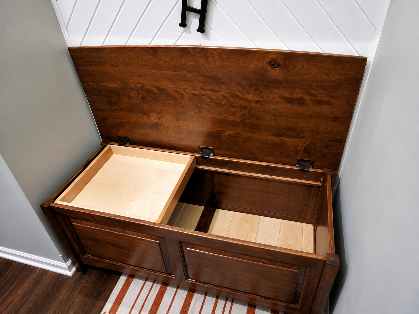 Open wooden storage bench with inside compartments, positioned against a corner wall with a decorative pattern, on a wooden floor with a striped rug nearby.