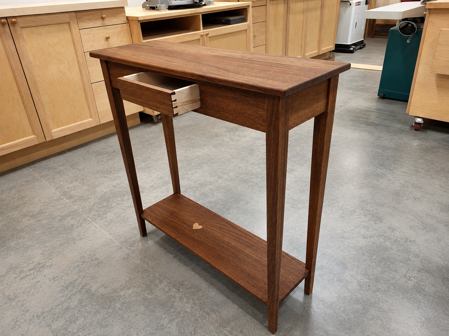 A wooden side table with a small drawer and a lower shelf, situated in a woodworking shop.