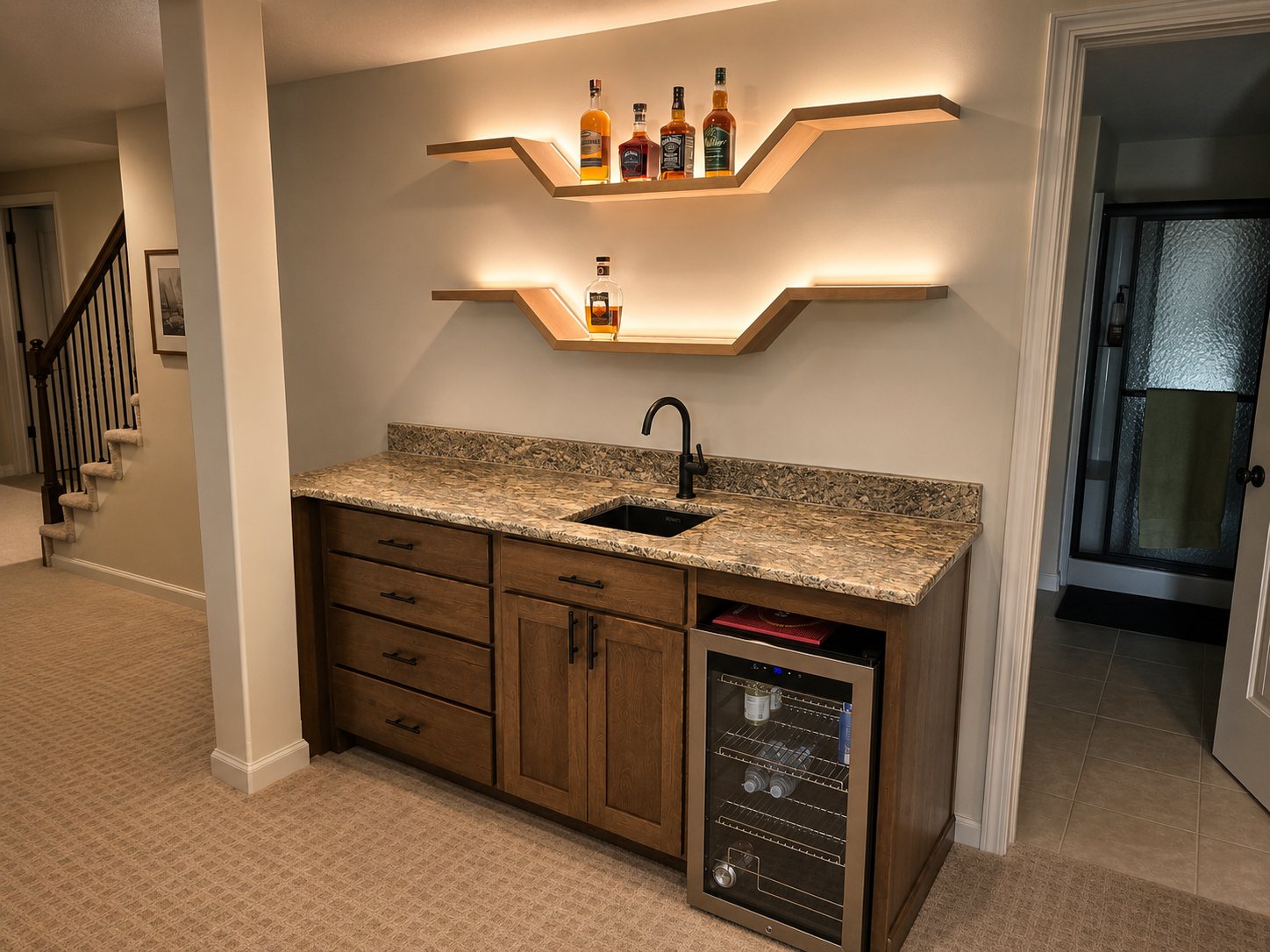 Kitchen area with wooden cabinets, granite countertop, a black sink, and a built-in mini fridge, with two modern zigzag wooden shelves above holding liquor bottles, lit with warm backlighting.