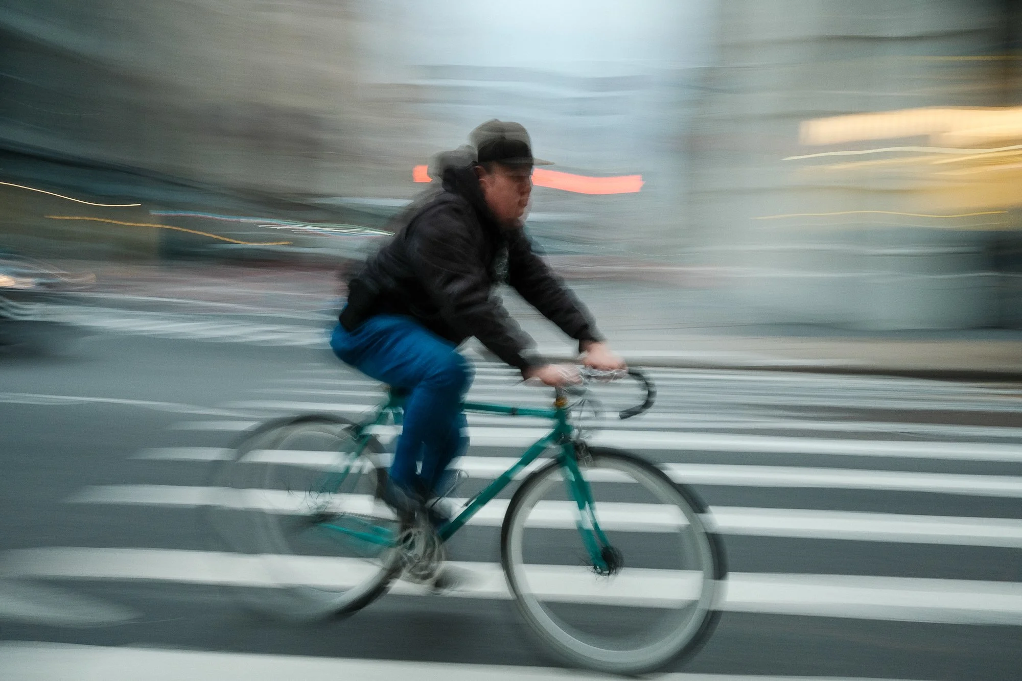 A person riding a bicycle across a zebra crossing in motion, with a blurred city background. The person is wearing a black jacket, gray cap, and glasses.