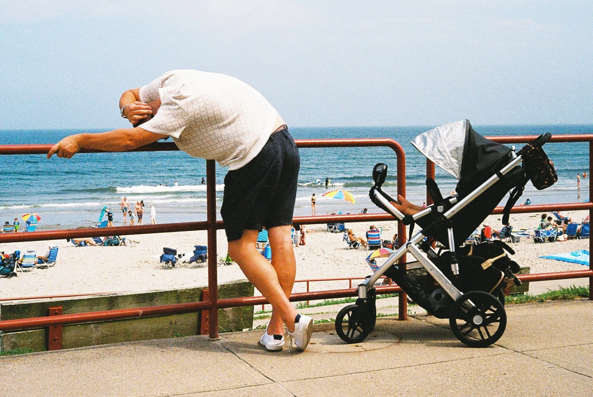 A man leaning over a red railing at the beach, with a stroller nearby. The beach is crowded with people, umbrellas, and chairs, and the ocean waves are visible in the background.