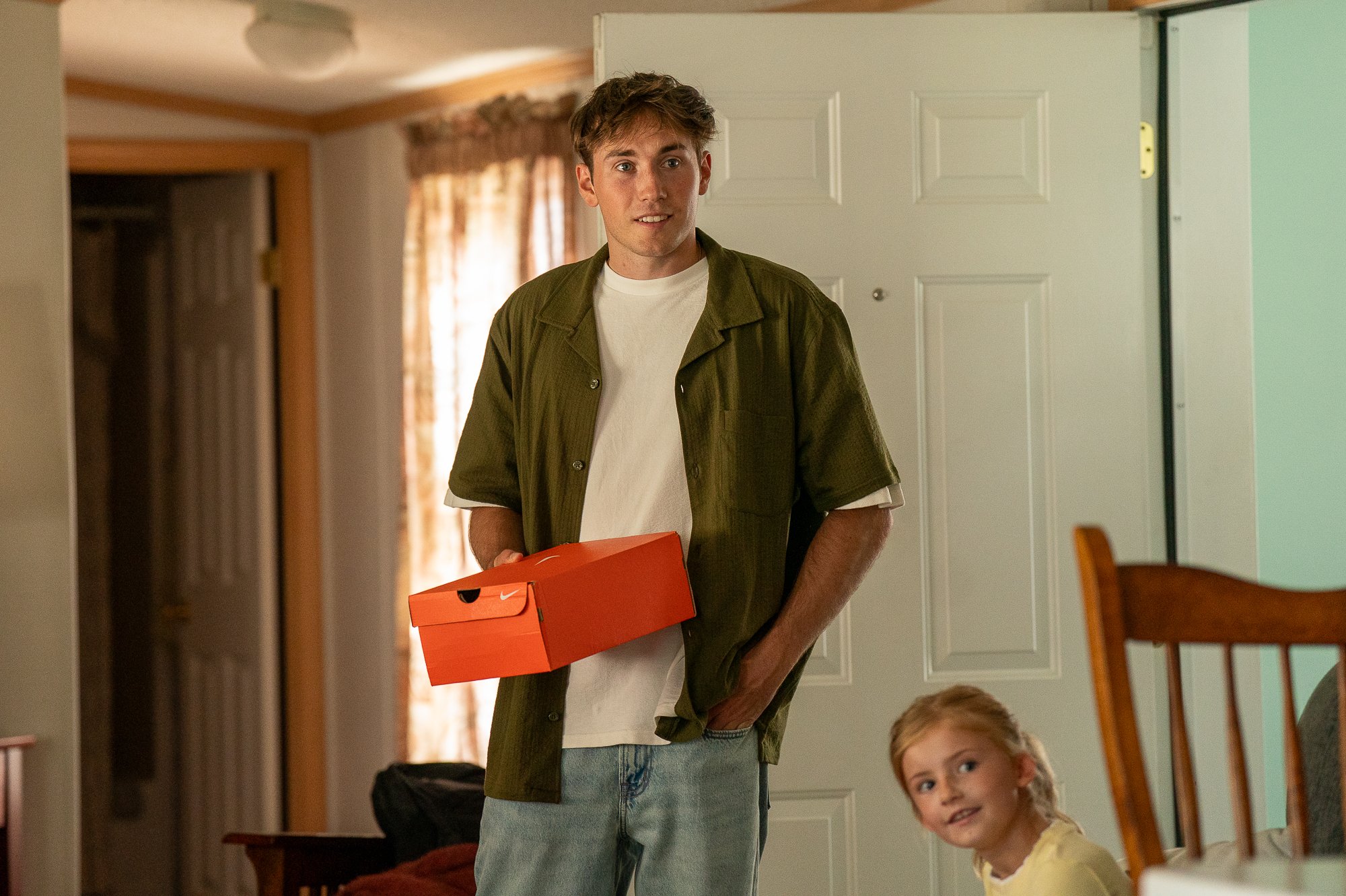 A young man wearing a green shirt and jeans holding an orange Nike shoebox inside a house, with a young girl sitting on the floor nearby. There are curtains and a door in the background.