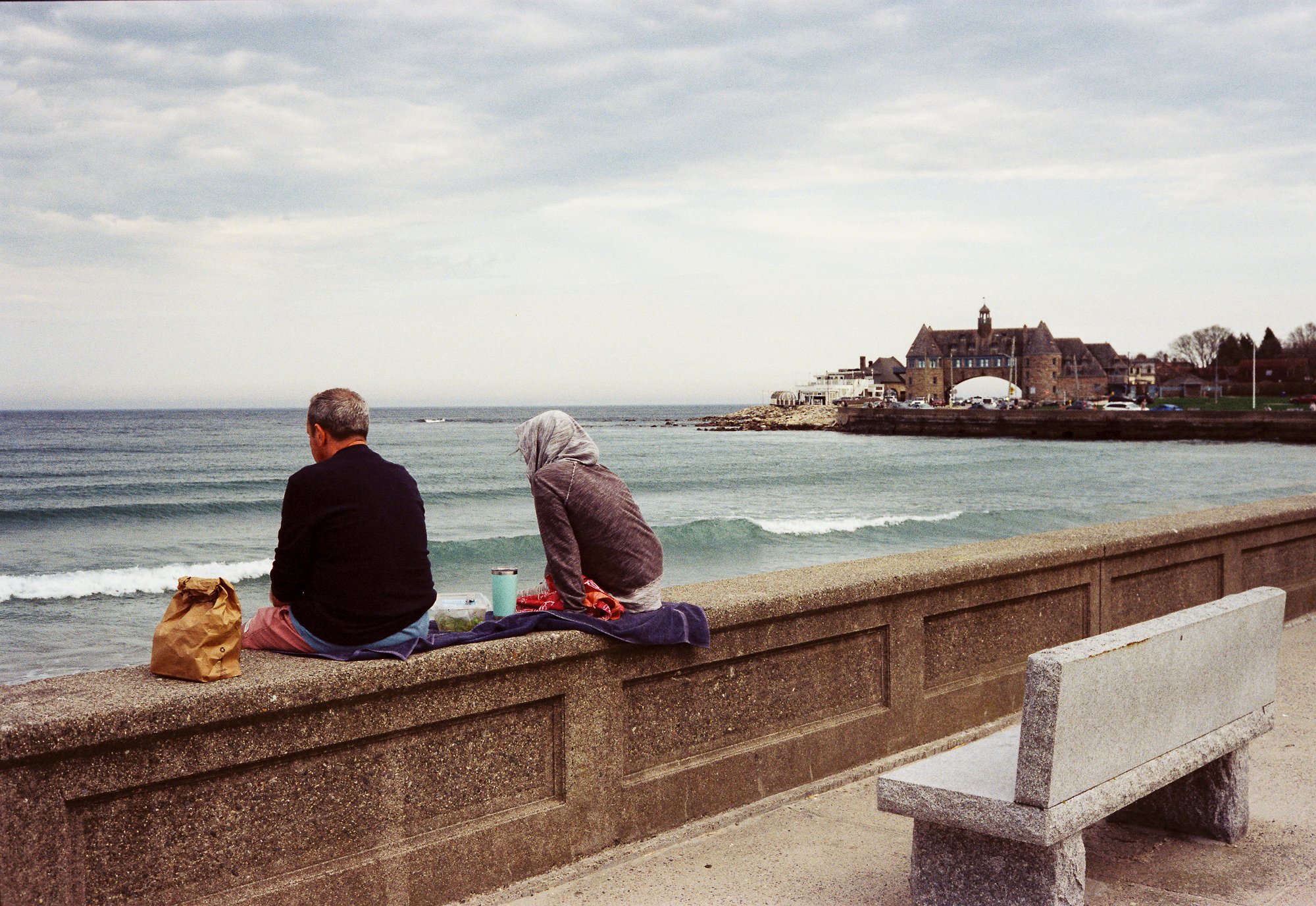Two people sitting on a concrete seawall, facing the ocean, with a cloudy sky overhead. One person has gray hair and a dark jacket, the other person has a head scarf and a gray sweater. There are items like a paper bag, thermos, and bags beside them.