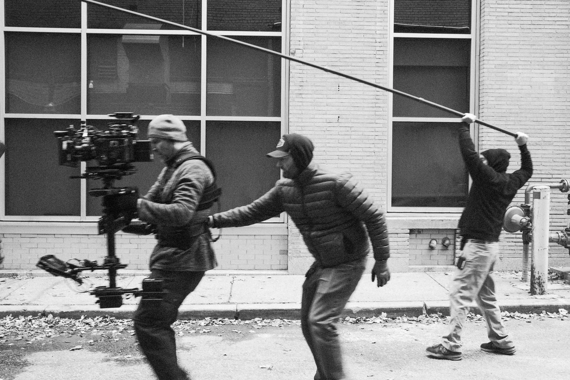 Three people walking along a sidewalk in front of a brick building with large windows. One person is operating a camera on a stabilizer, another is walking behind, and a third person in the background is holding a boom microphone.