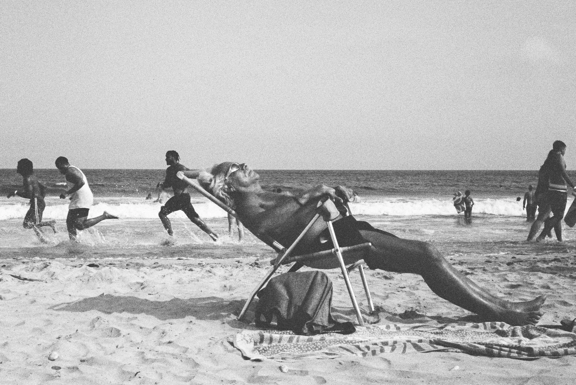 Black and white photo of people at the beach, some running and playing in the water, with a large seashell in the foreground.
