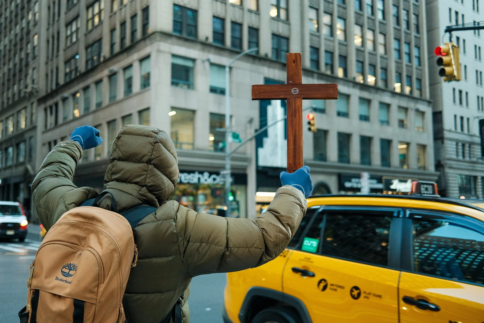 A person wearing a brown jacket and blue gloves holds a wooden cross in front of a city street scene with yellow taxis, a traffic light, and tall buildings.