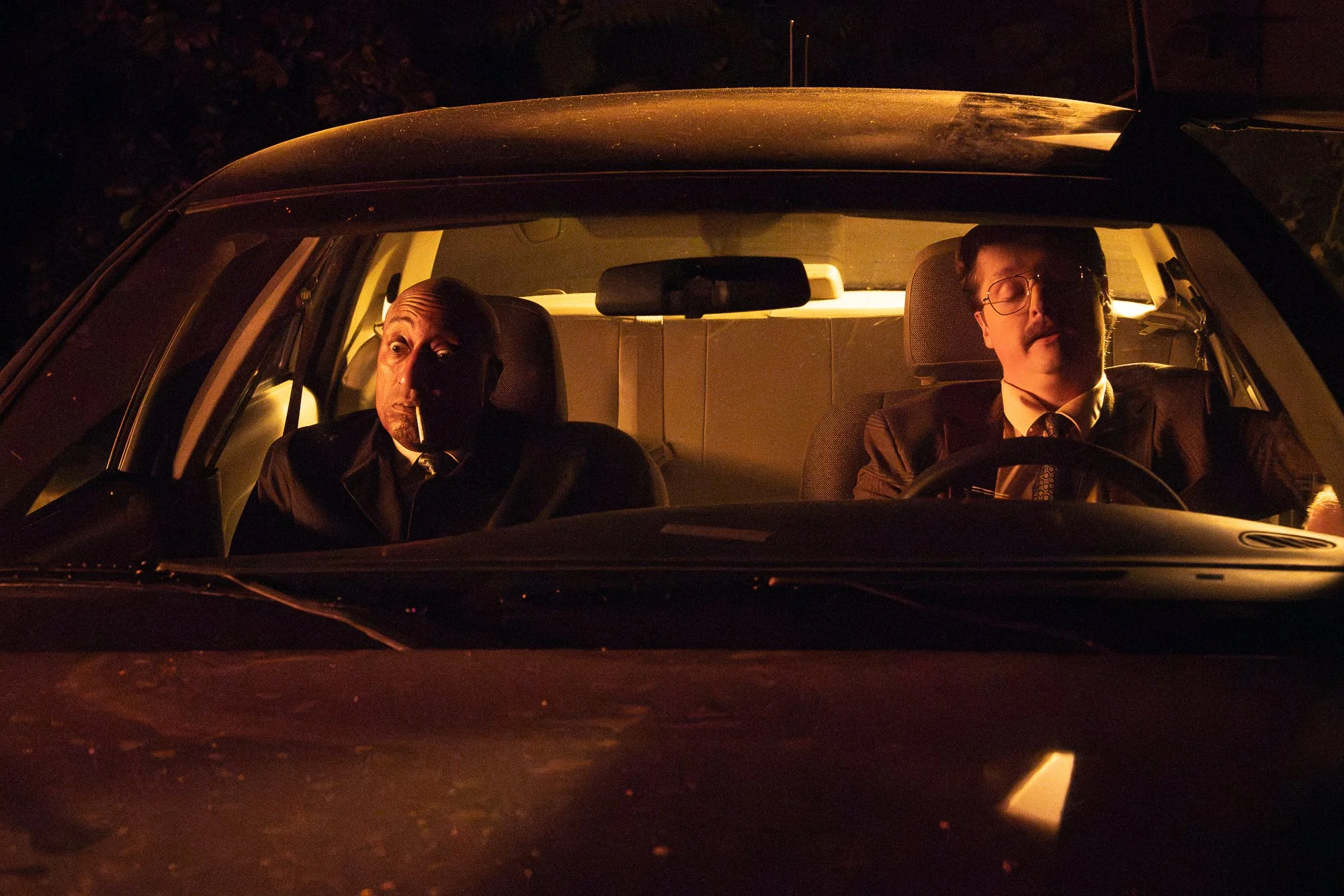 Two men sitting inside a car at night, illuminated by the car's interior light, with one smoking a cigarette and the other wearing glasses.