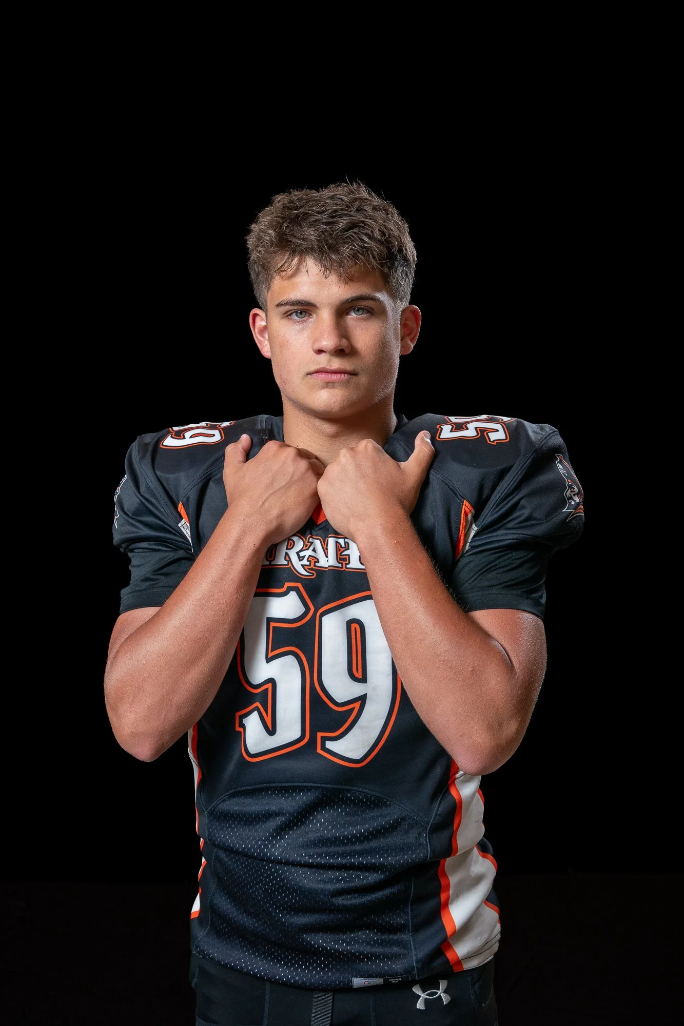 A young male football player with short brown hair and blue eyes posing against a black background. He is wearing a dark football jersey with orange and white accents, the number 59 prominently displayed on the front, and is holding his hands near hi