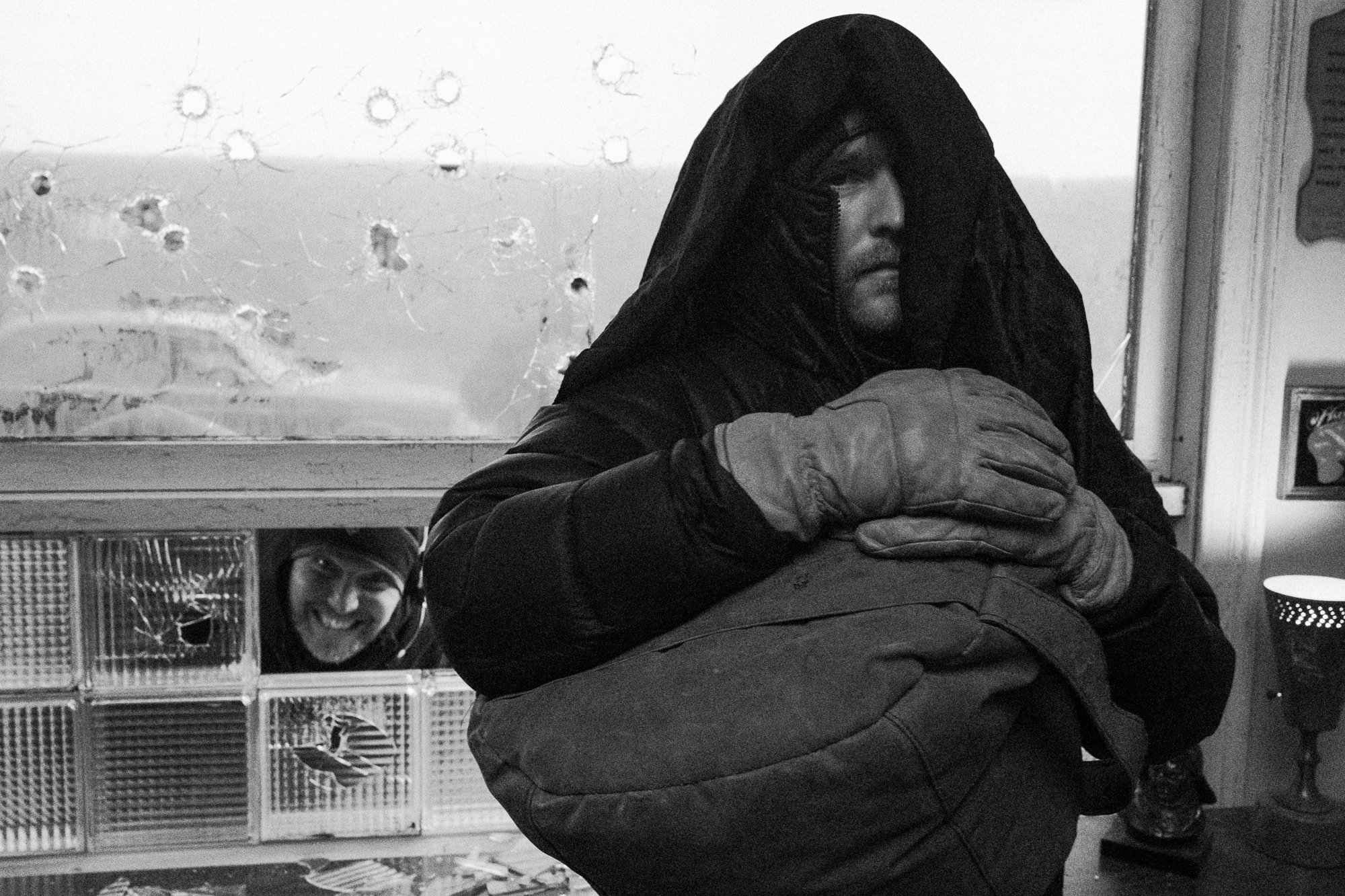 Black and white photo of a man with a hooded jacket and gloves, looking to the side, with a woman smiling and wearing glasses in the background. The setting appears to be indoors with patterned decor.