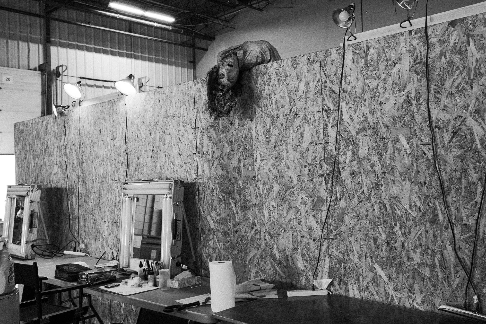 A workshop or studio with a wooden wall and a large, realistic lion mask or sculpture hanging on the wall. There are work tables with various tools and equipment, including 3D printers, in the foreground. Several professional studio lights are direct