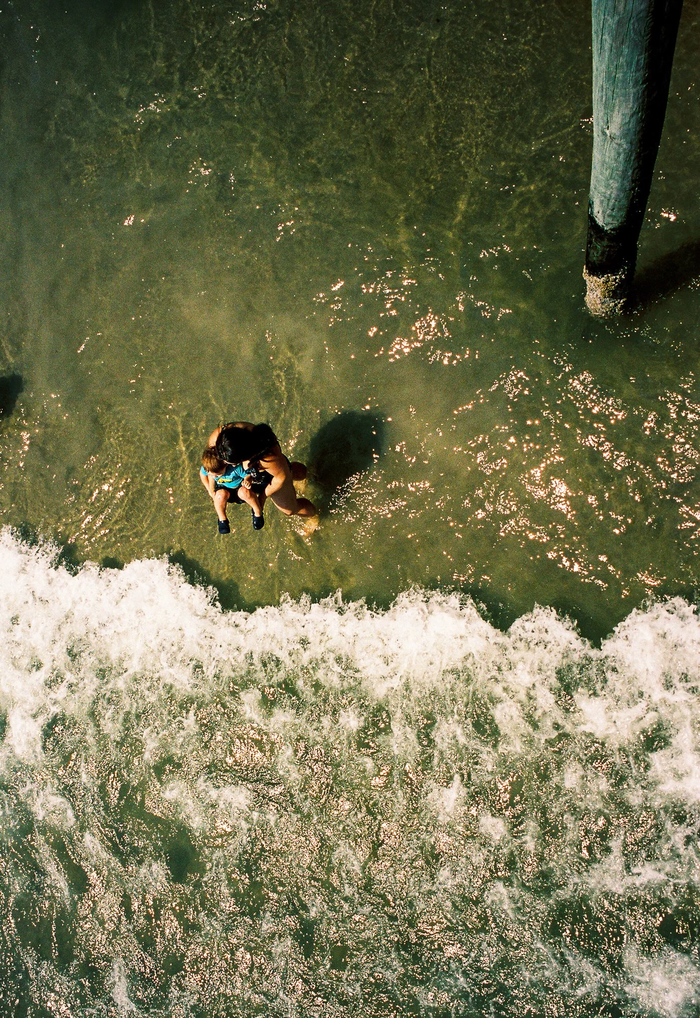 A bird's eye view of a person standing in shallow water near a wooden pole, with waves crashing on the shore at the bottom of the image.