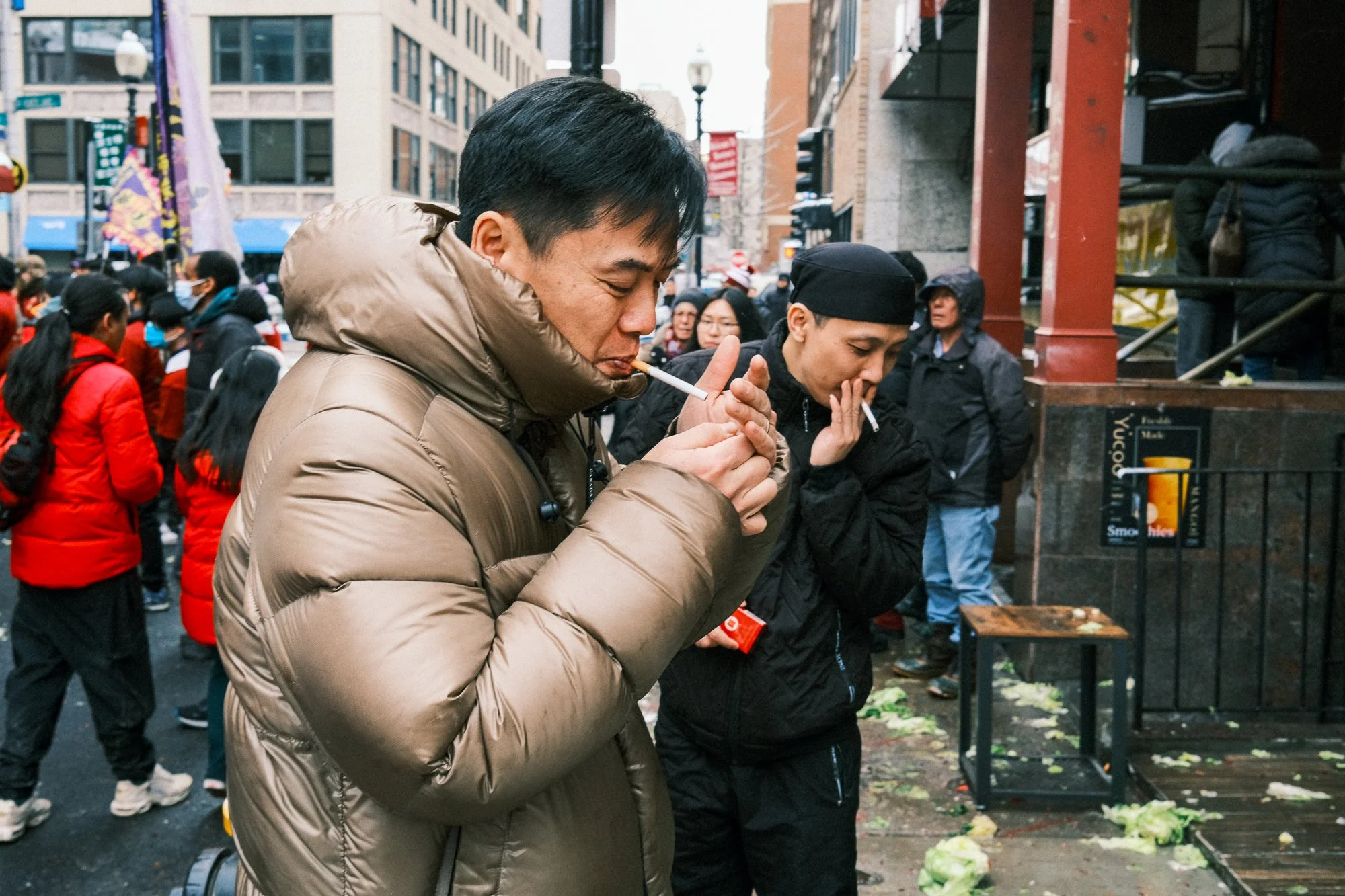 Two people engaged in a memorial or vigil, lighting candles or incense in a city street, surrounded by a crowd of onlookers.