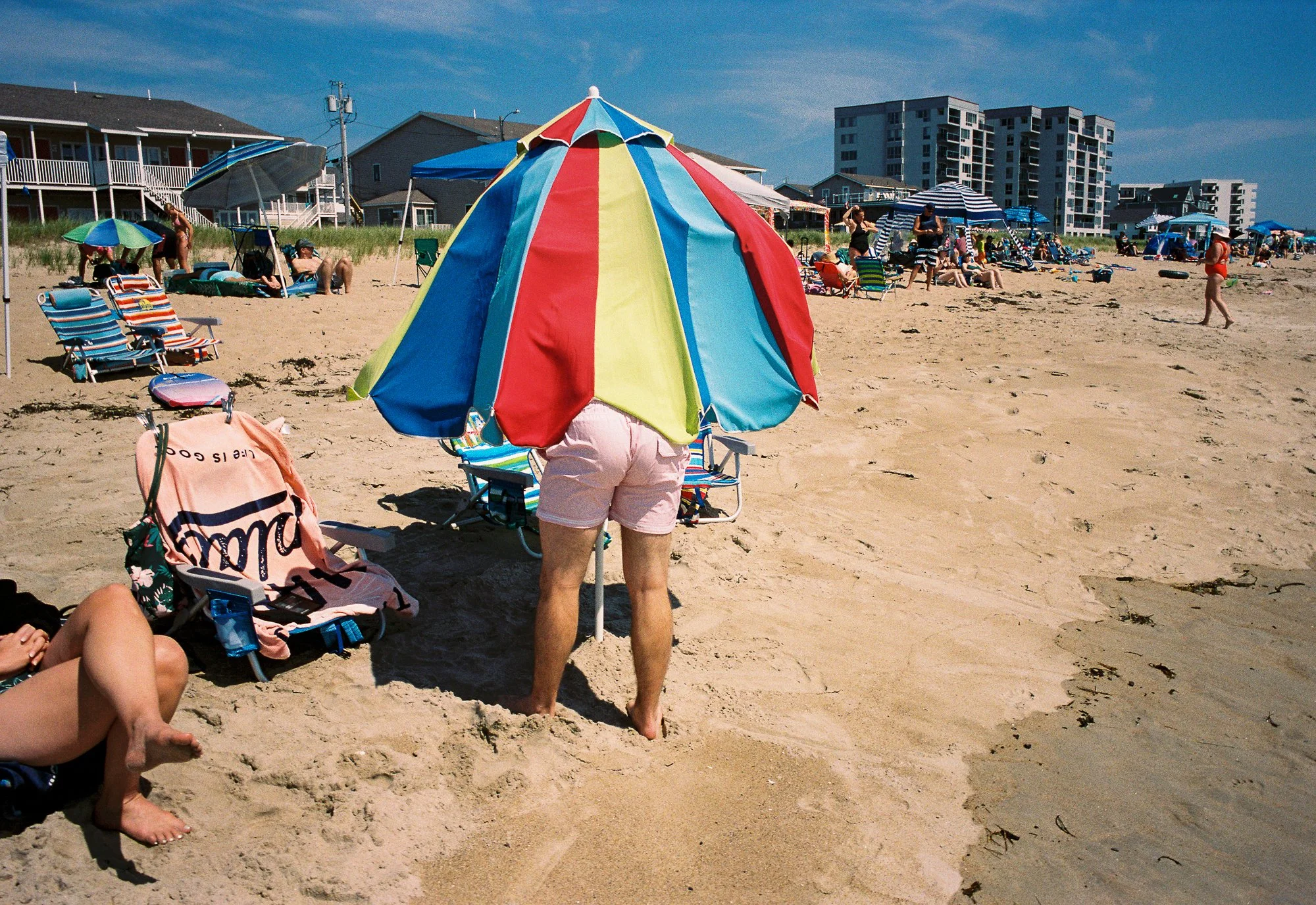 People relaxing and socializing on a sandy beach with umbrellas and beach chairs, with residential buildings in the background under a clear blue sky.