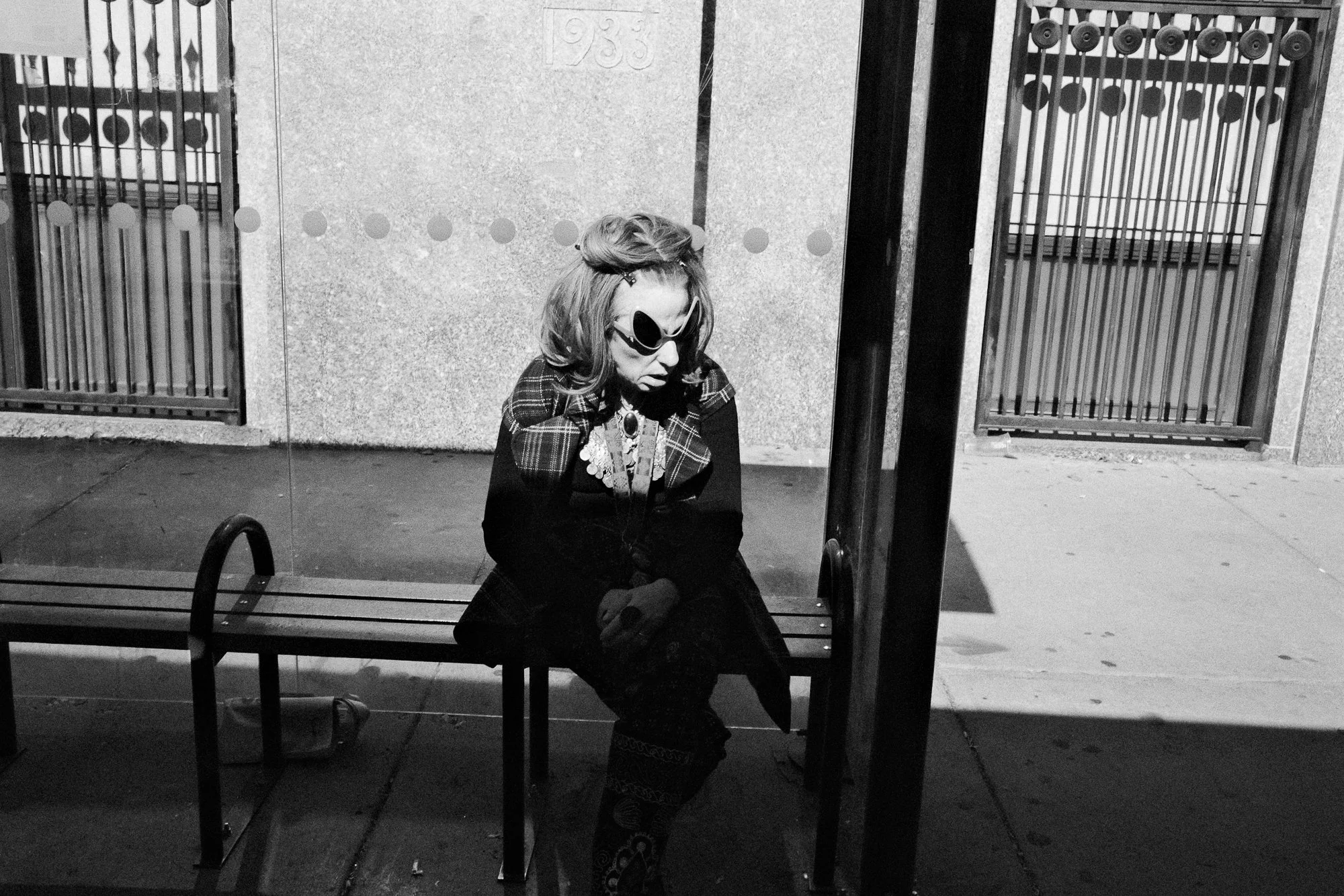 A woman with wavy hair wearing sunglasses, a plaid jacket, and floral pants sitting on a bench at night, looking downward, with a large bag on the ground beside her, reflected in a glass window.