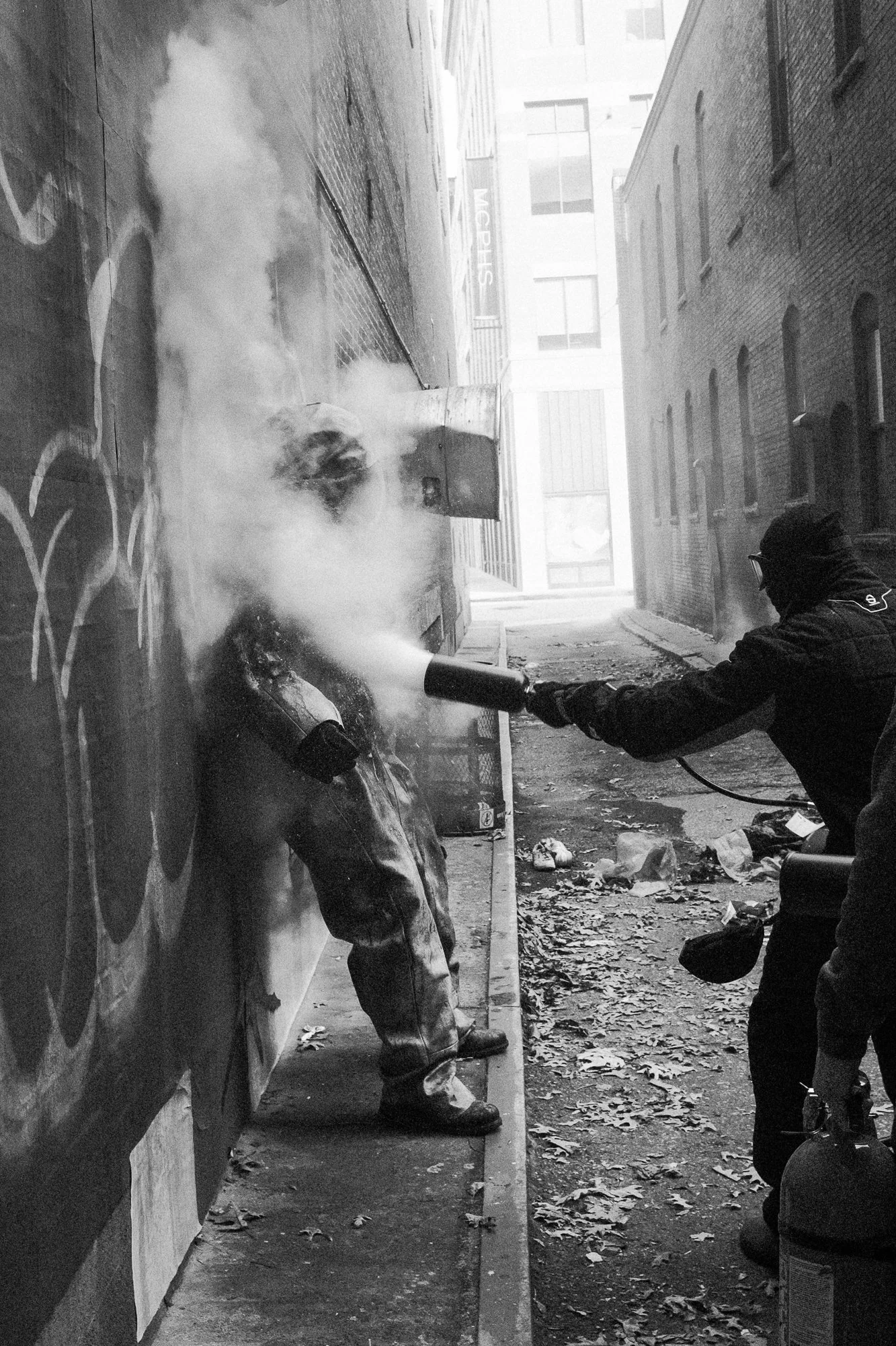 A person wearing a hooded jacket is using a fire extinguisher to put out a fire on a car in an alleyway, with smoke billowing upwards. The alley has graffiti on one wall and debris scattered on the ground, with tall buildings visible in the backgroun