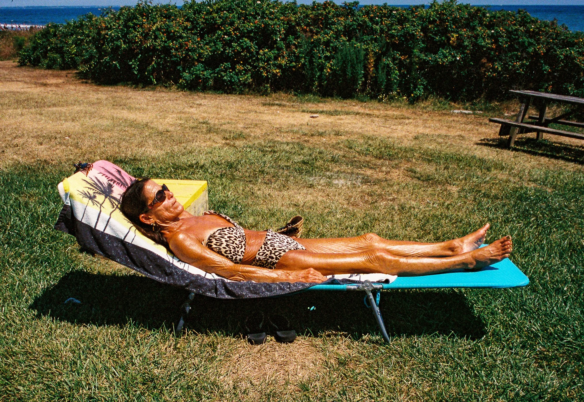An elderly woman with dark skin and sunglasses is lying on a lounge chair on a grassy area near the beach, wearing a leopard print swimsuit, with a colorful beach towel under her head and a blue mat underneath her legs. She is relaxing in the sun wit
