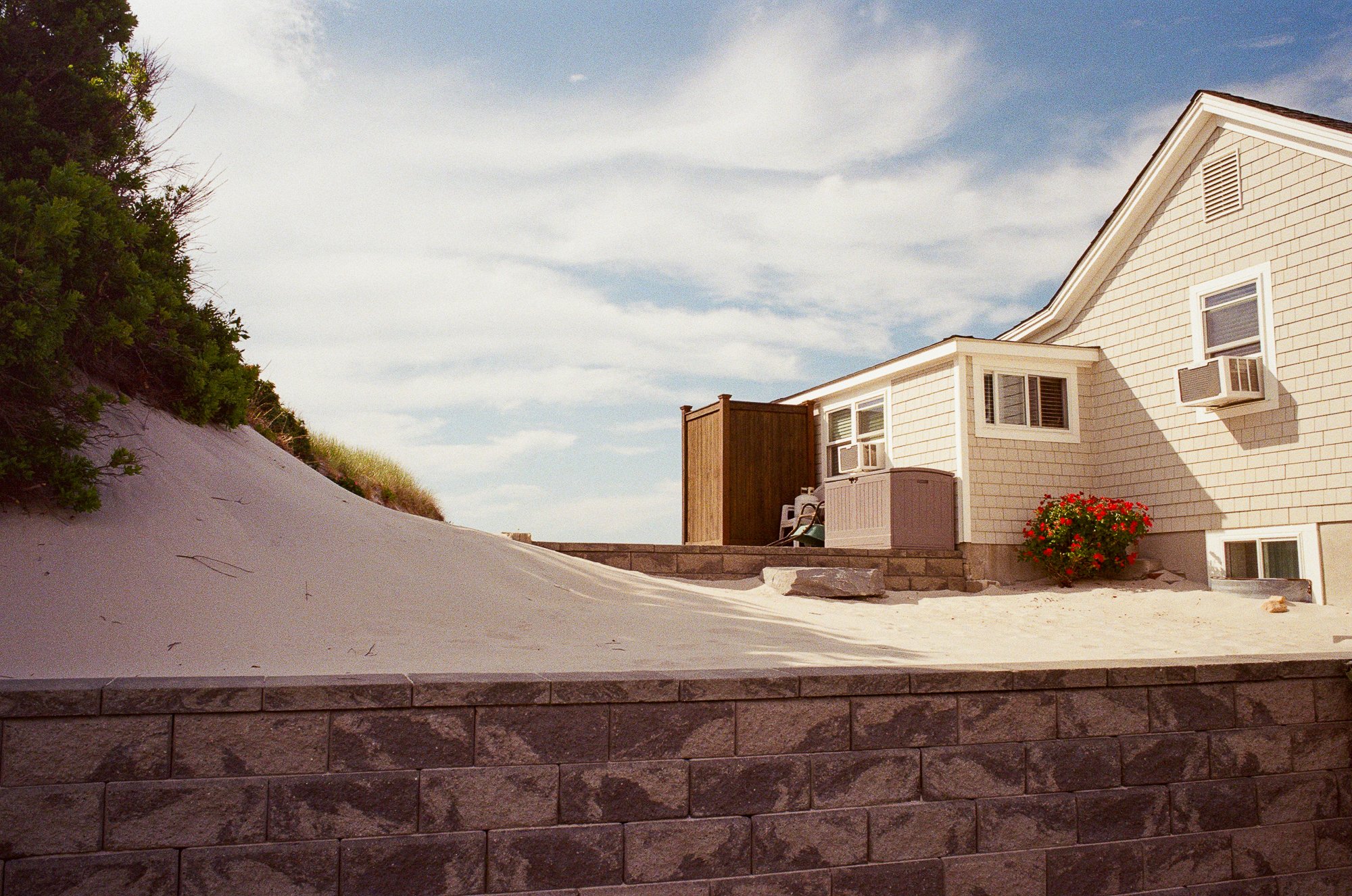 A residential backyard with sand, a brick wall, a house with septic windows and air conditioning units, a wooden privacy screen, outdoor furniture, a potted plant with red flowers, and a sand dune with green bushes and trees.
