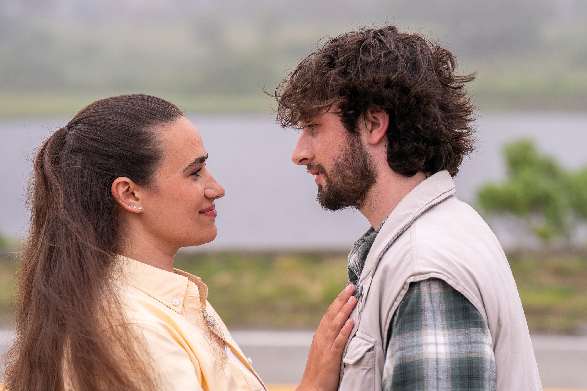 A man and a woman face each other closely outdoors, with the woman gently touching the man's chest. They are looking into each other's eyes and smiling softly. The background appears to be a natural landscape with water and greenery.
