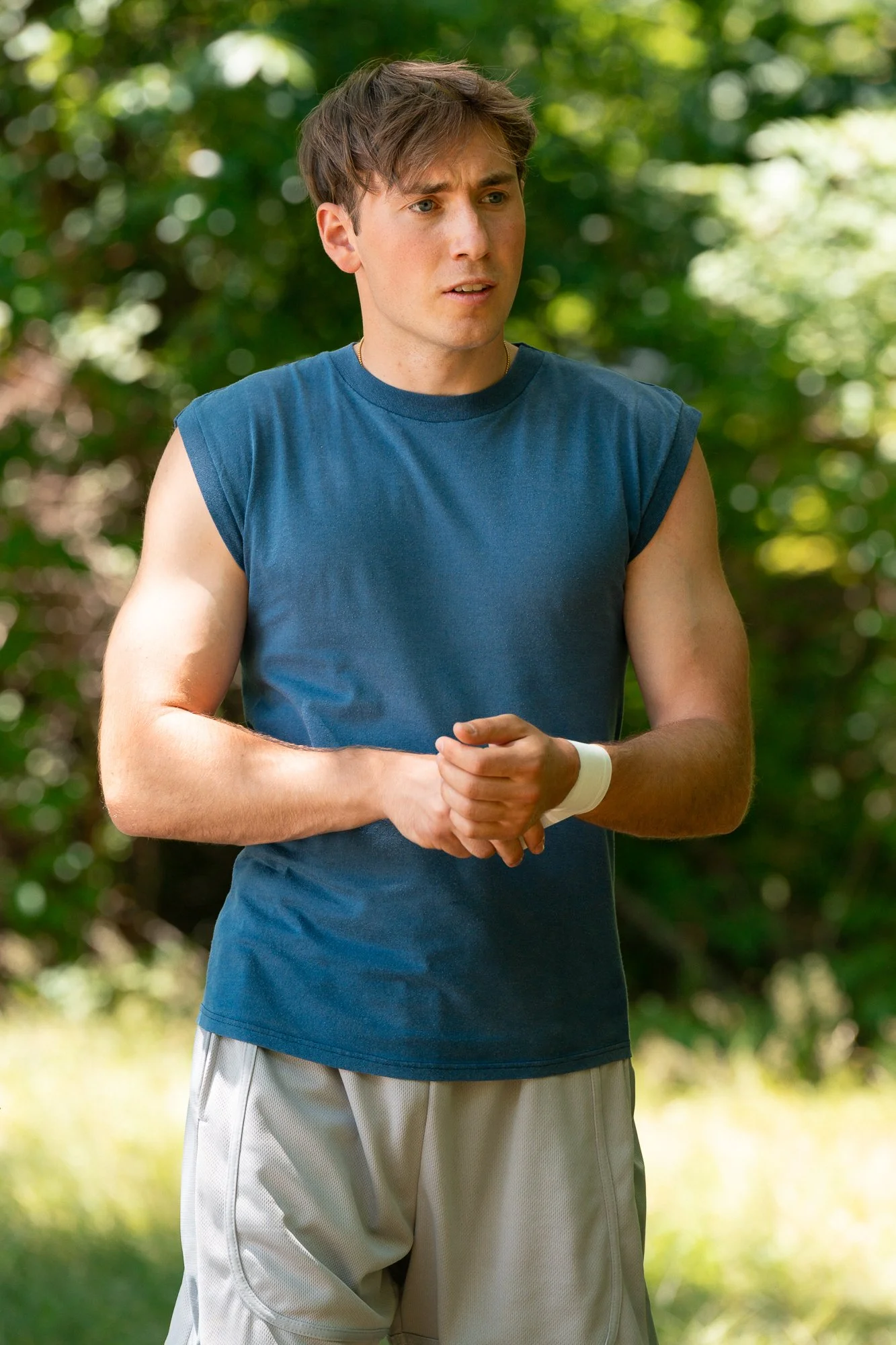A young man wearing a sleeveless blue athletic shirt and light-colored shorts with a white wristband on his left wrist, outdoors in a wooded area with greenery in the background.
