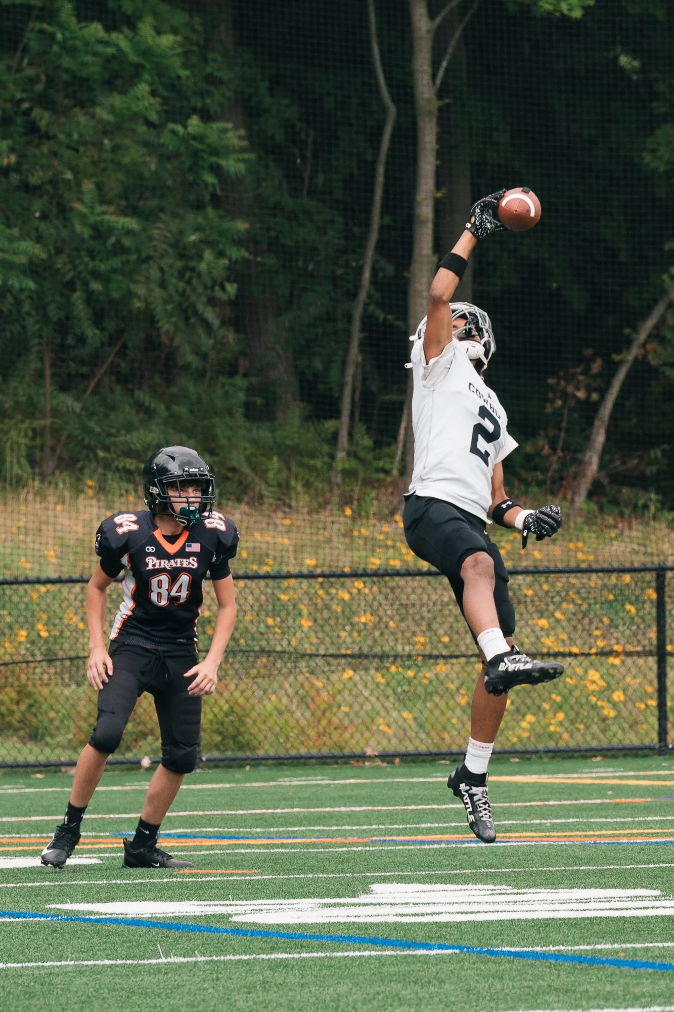 Young football players on field, one jumping to catch football, other standing watching, both in football gear, trees in background.