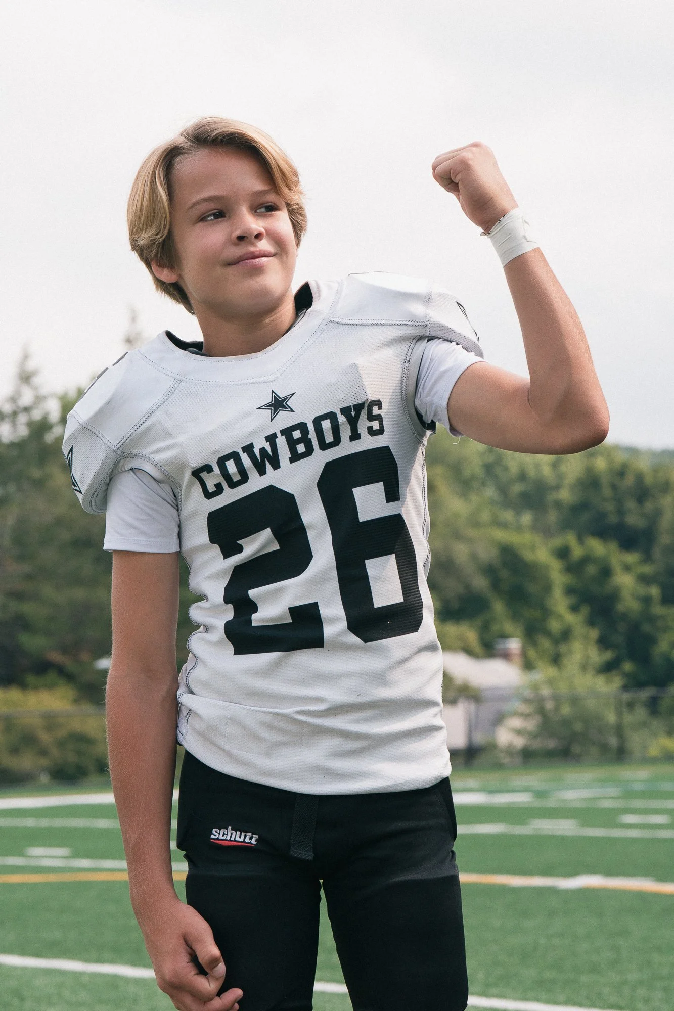 A young boy in a football uniform flexing his arm muscles on an outdoor football field.